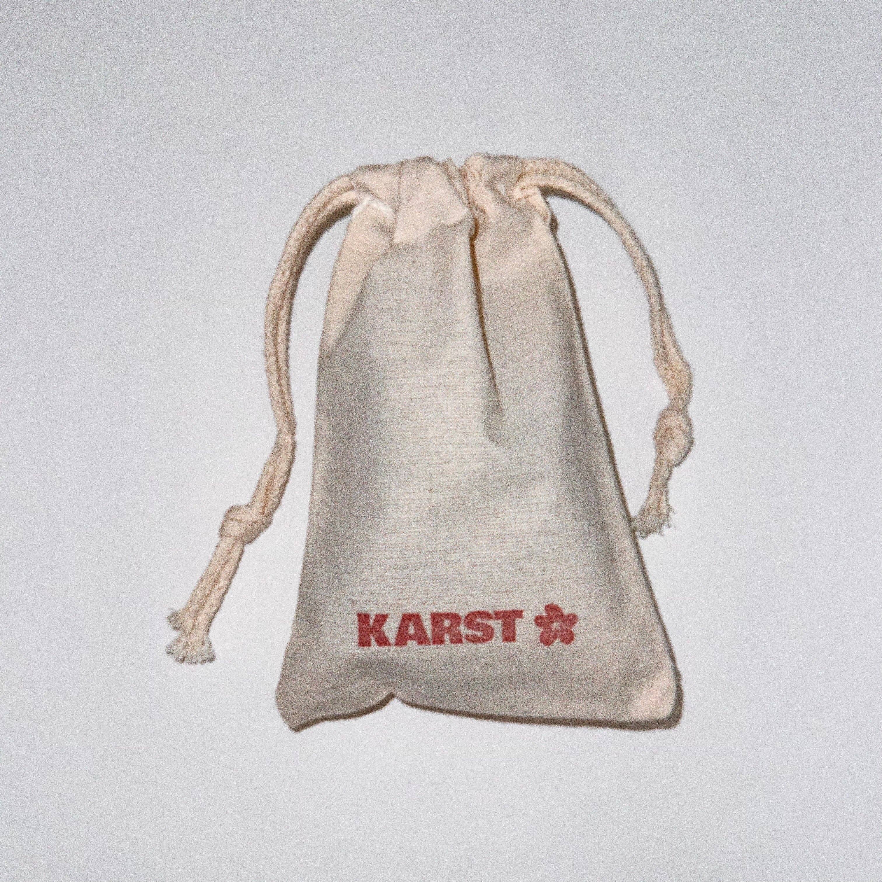 A small beige drawstring pouch with KARST and a red flower logo on the front, perfect for storing your Karst Essentials Slim Hair Claw Clip — Olive, shown on a plain white background.