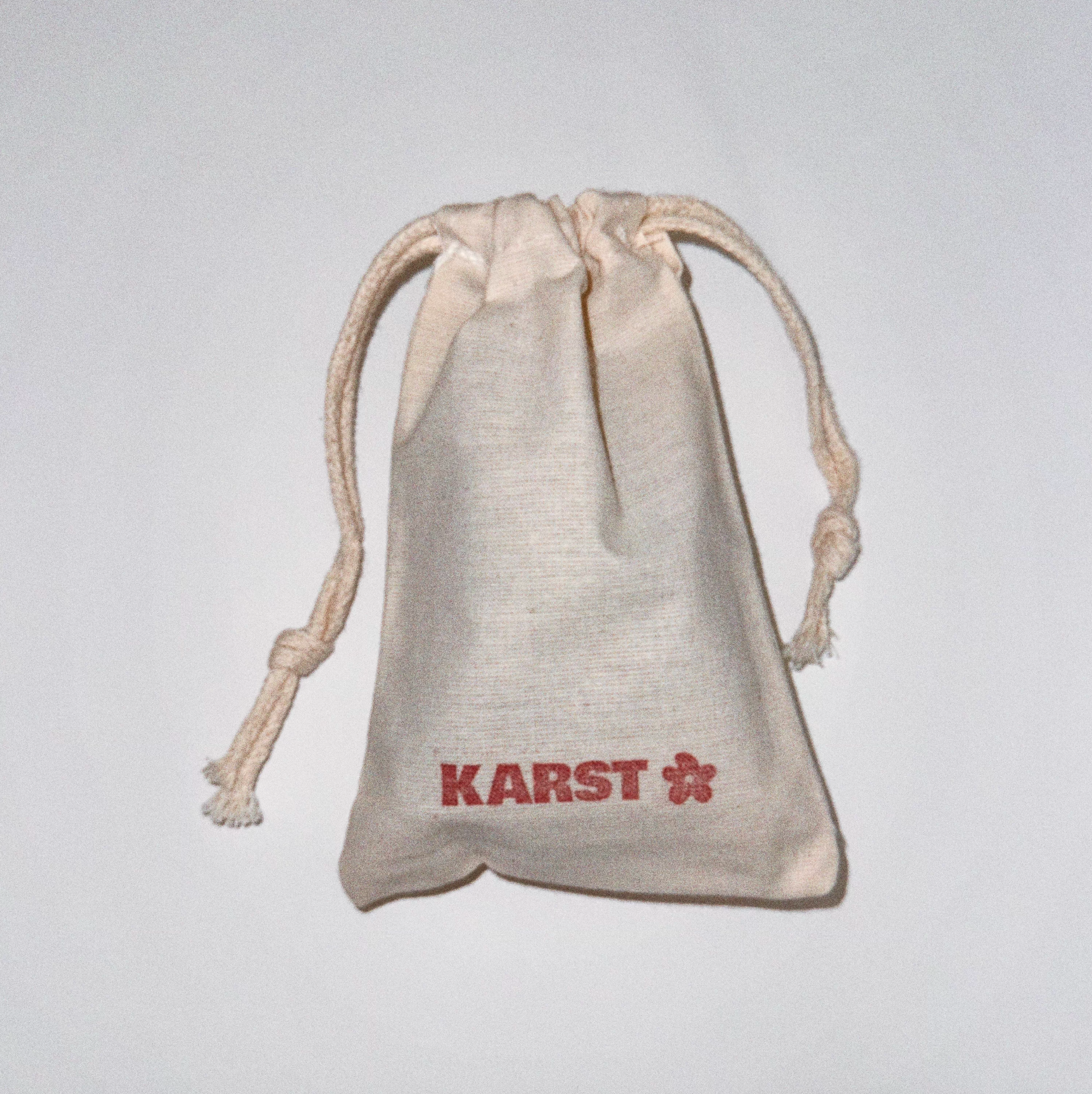 A small beige drawstring pouch with "Karst Essentials" and a red flower symbol on the front, ideal for storing your SuperClip Hair Claw — Mist: No Charm Eyelet (Standard), shown against a plain white background.