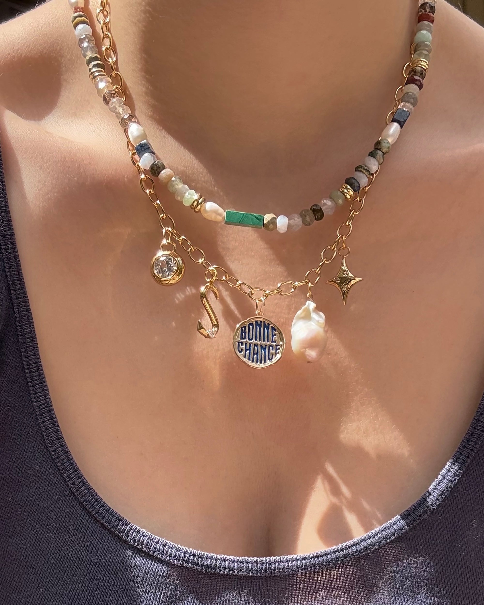 A close-up shows a person wearing the Mod + Jo Andy Gemstone Necklace: layered with a beaded choker and 14k gold-plated chain, featuring five unique pendants over a dark tank—genuine gemstones, hook, “BONNE CHANCE” circle, baroque pearl, and star.