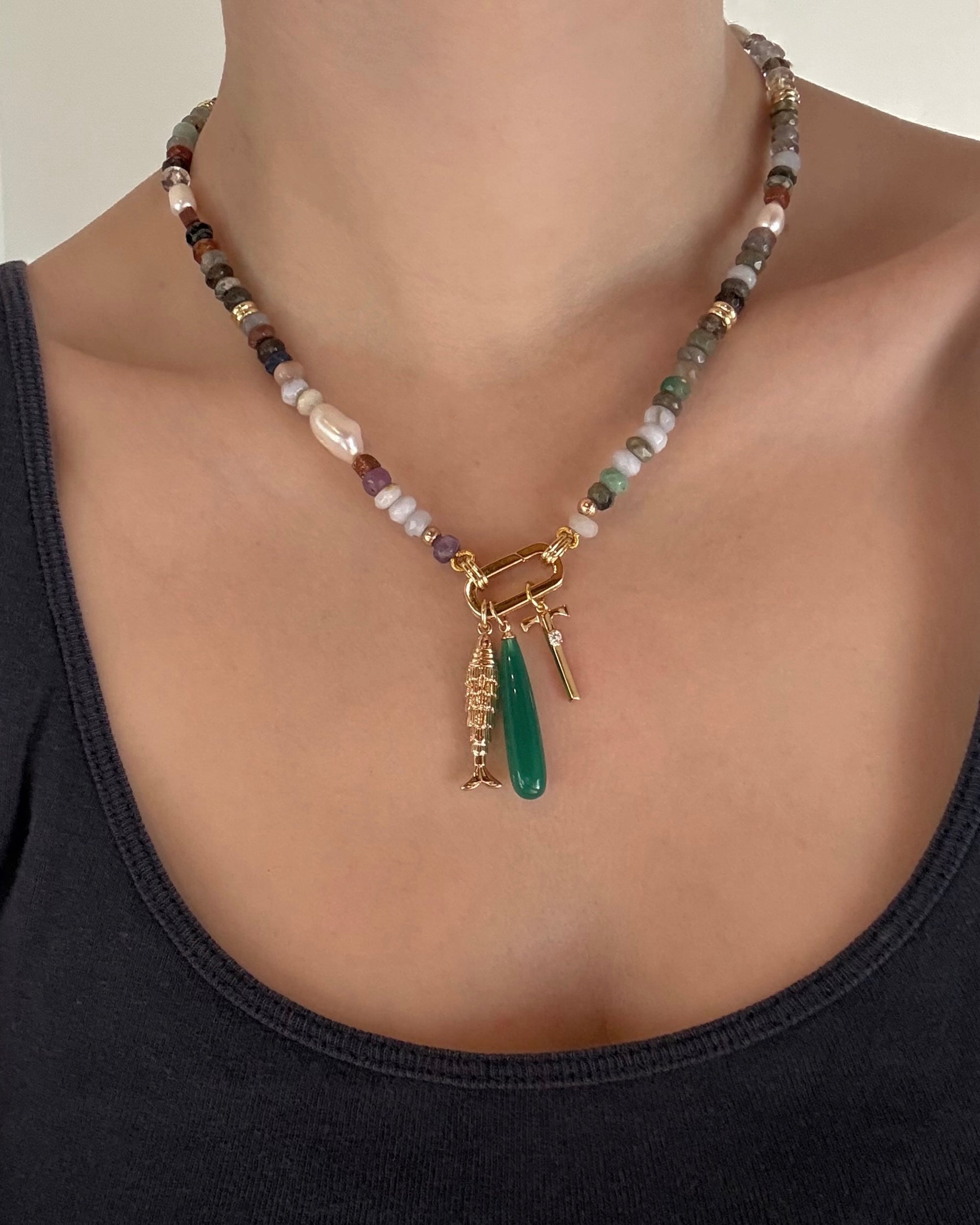 A person wears the Mod + Jo Andy Gemstone Enhancer Chain, a beaded necklace with genuine gemstones and three gold charms—a green pendant, a textured gold charm, and a small gold bar—layered over a dark tank top.