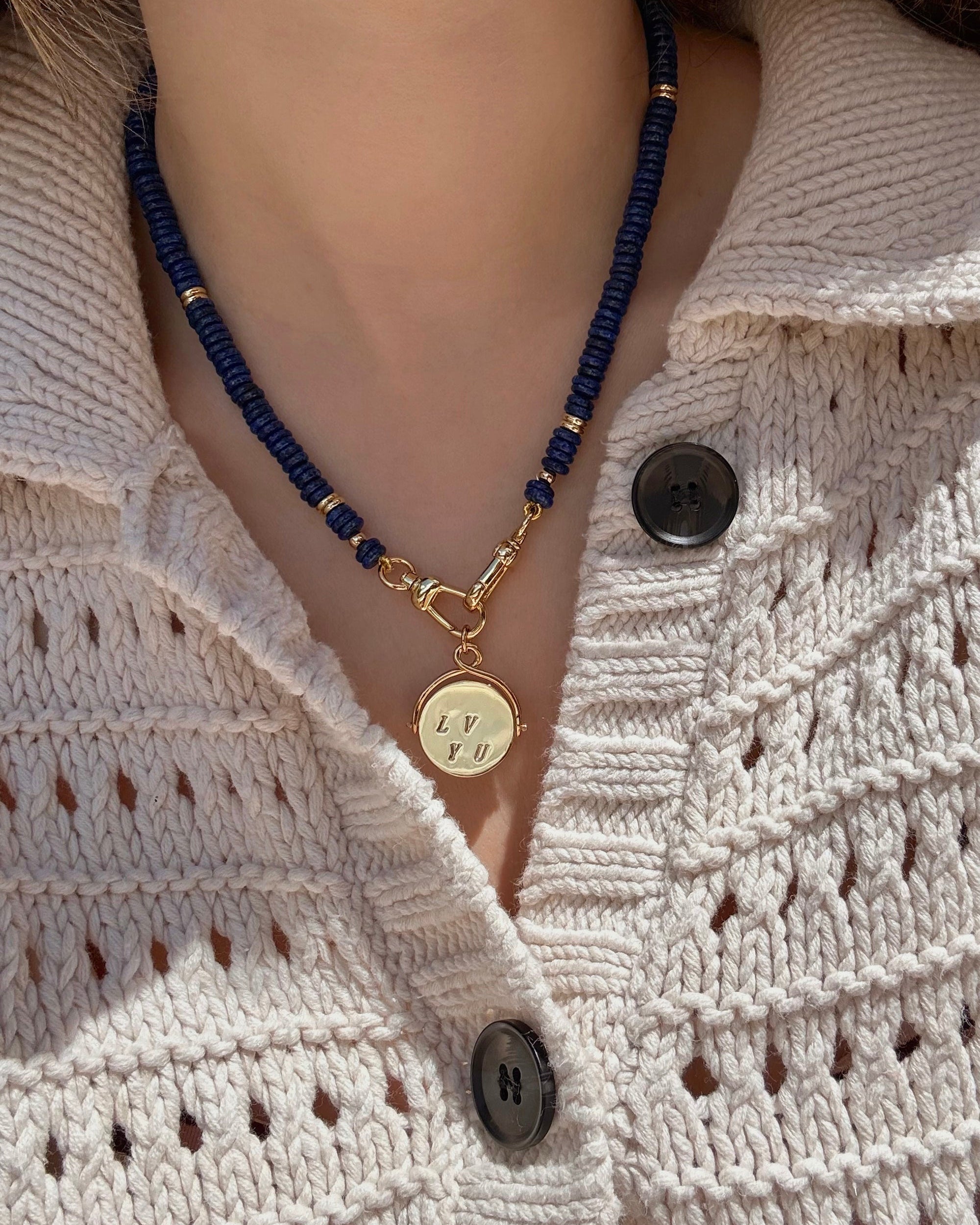 A person in a cream knit sweater with black buttons and a dark blue bead necklace, featuring the Mod + Jo Bella "I Love You" Spinner Charm in 14k gold as playful fidget jewelry.