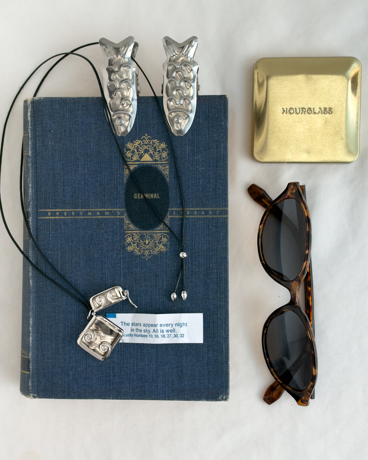 A blue hardcover book sits next to a silver Sybil Vesta Case Charm on a cord, two fish-shaped hair clips, a gold compact, and tortoiseshell sunglasses.