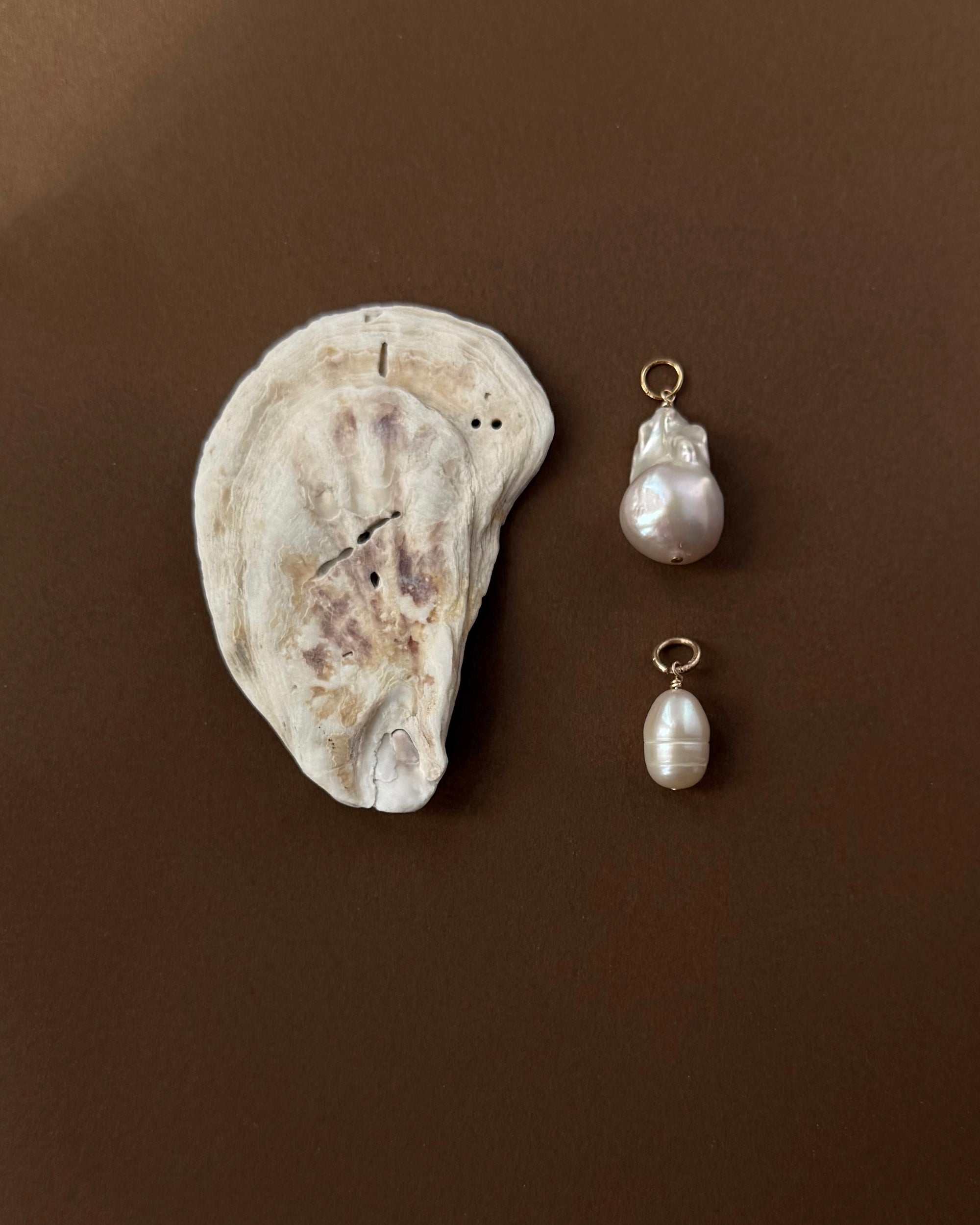 A flat, textured oyster shell rests on a brown surface beside two Mod + Jo charms: the Jaci Baroque Pearl Charm with an irregular baroque pearl and another featuring a smooth, oval freshwater pearl—both 14k gold plated.