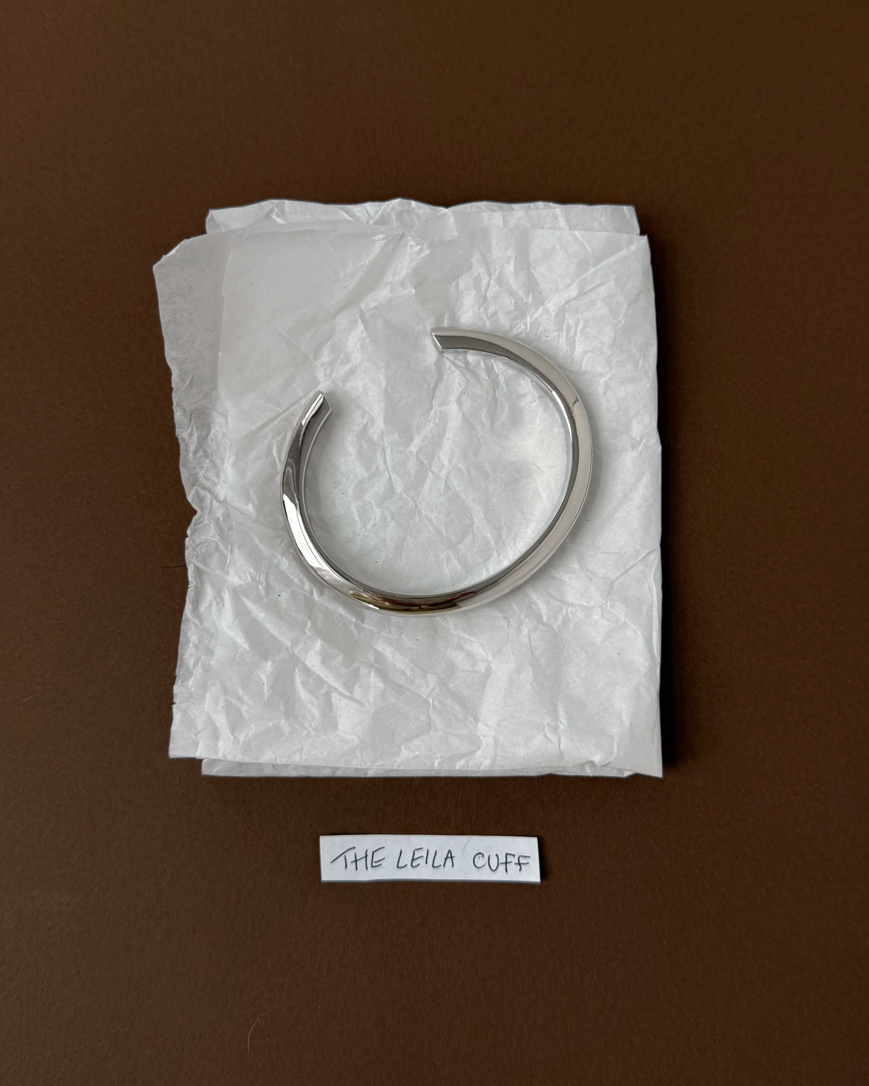 A sleek, rhodium-plated Leila Dome Cuff Bracelet by Mod + Jo rests on crumpled white tissue atop a brown surface, with a small label reading THE LEILA CUFF below.