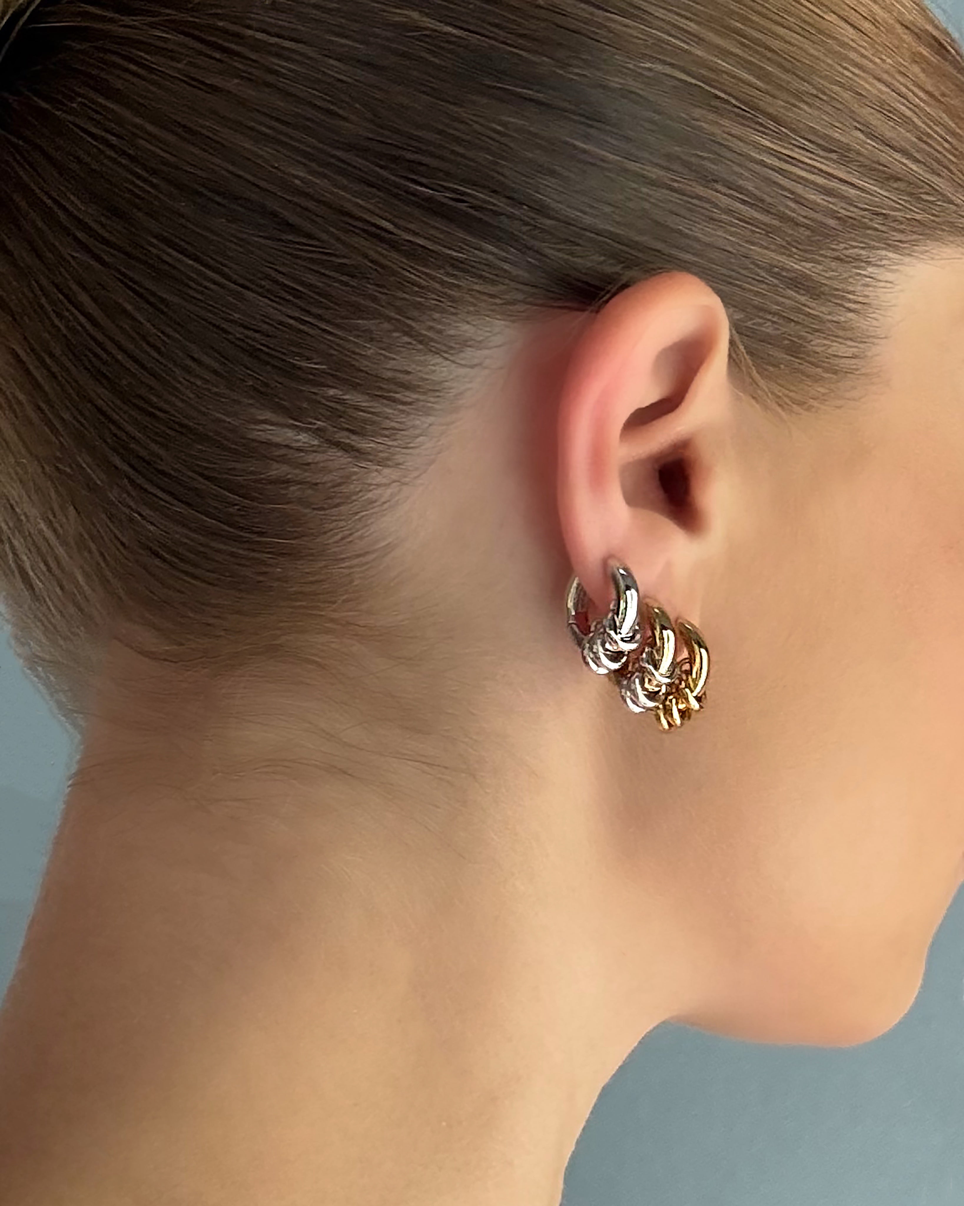 Close-up of a woman’s ear adorned with layered silver and gold plated huggie earrings, featuring the Mod + Jo Grande Levi Huggies—which are nickel and lead free—with her brown hair pulled back against a neutral background.