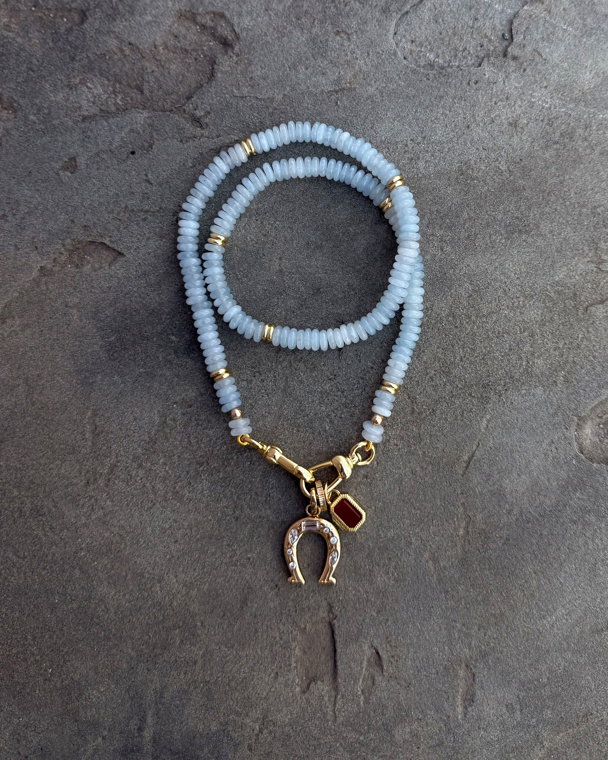 The Mod + Jo Grande Chantal Horseshoe Charm bracelet features pale blue beads, gold accents, a gold horseshoe charm with clear stones, and a red charm—shown on a textured gray surface for a touch of luck.