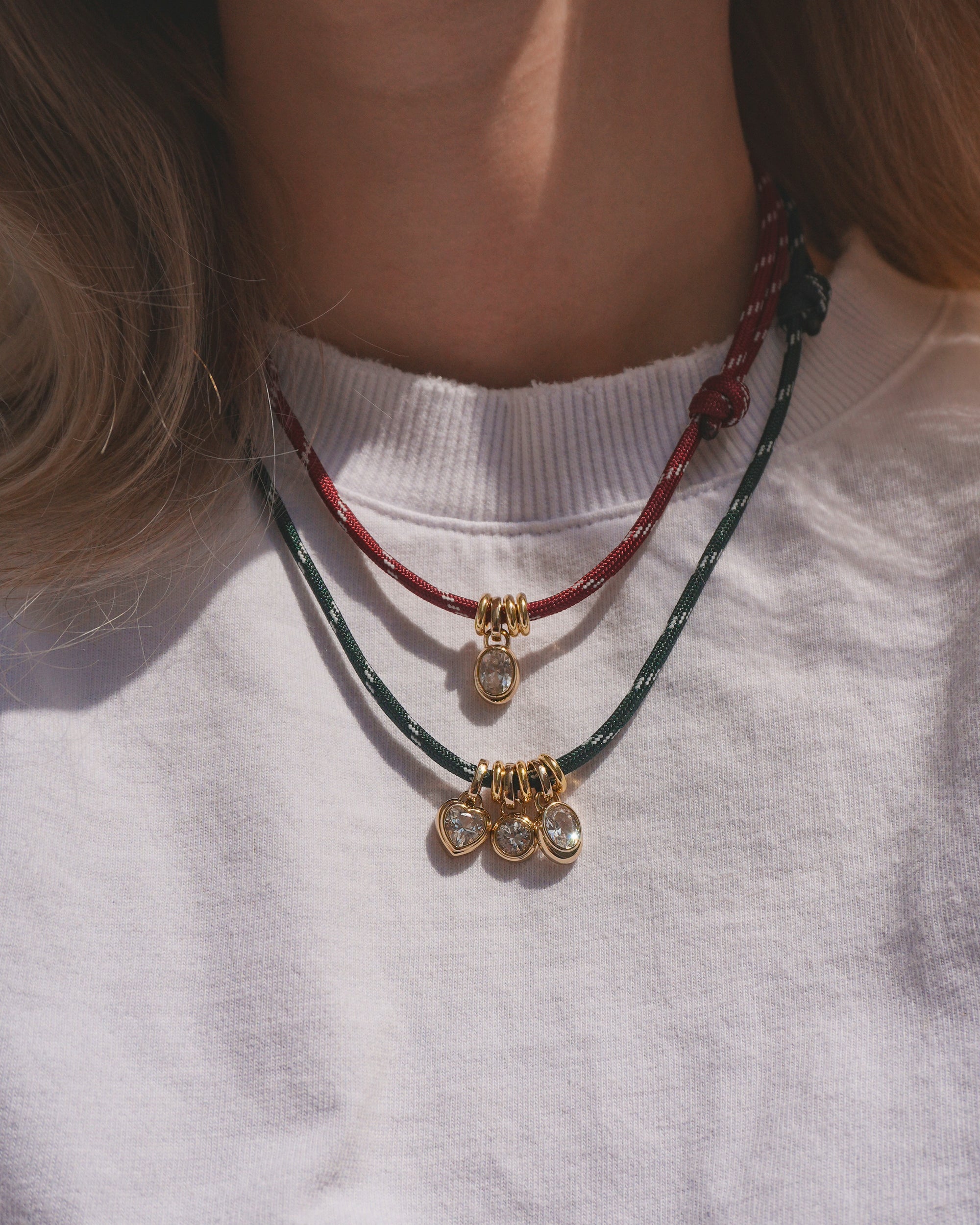 A person wearing a white shirt styled with two Mod + Jo Tommy Cord Necklaces: one in red with an adjustable design and a single gold and crystal pendant featuring gold-plated accents, and another in green paracord adorned with three gold and crystal pendants.