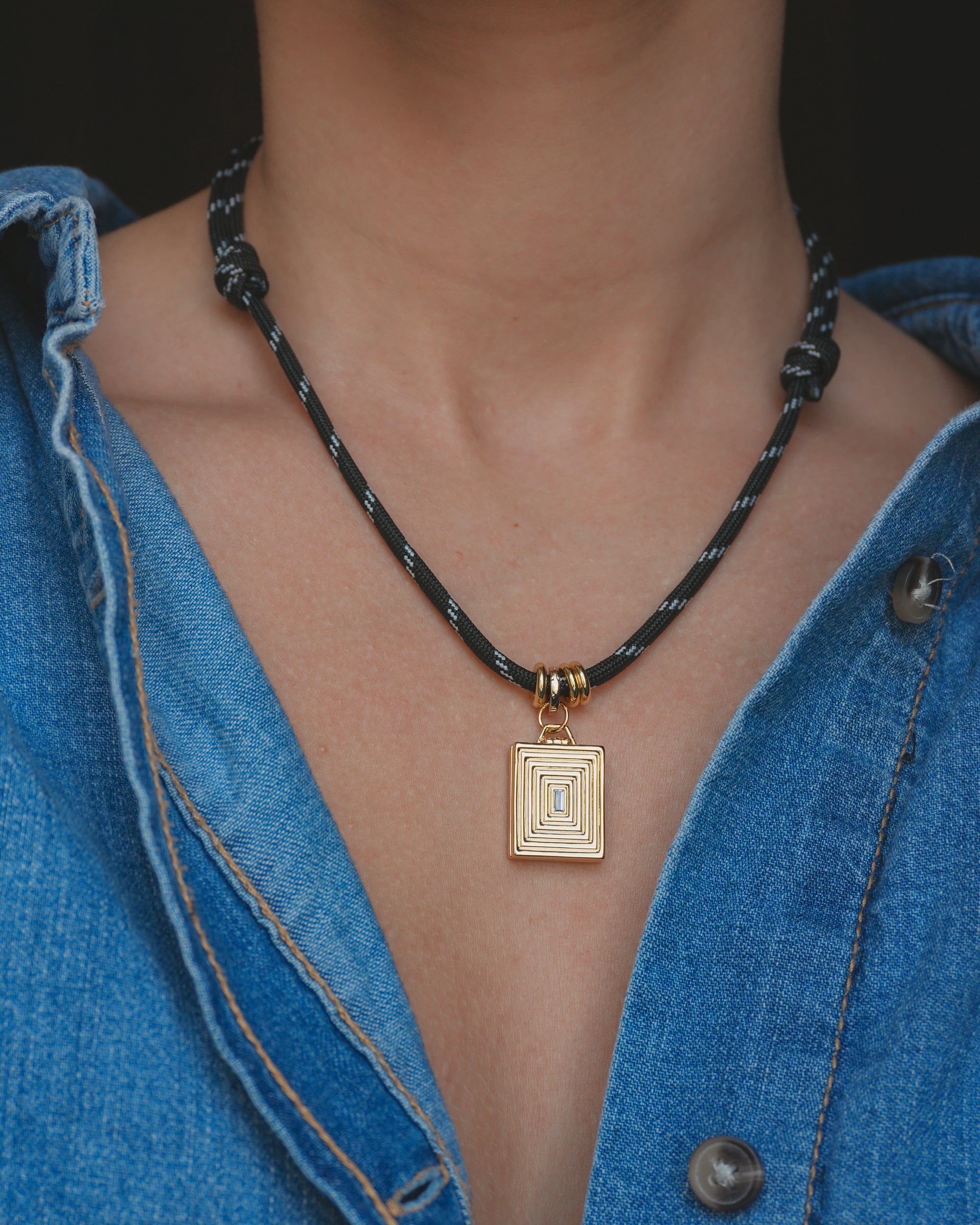 A person wearing a blue denim shirt with an open collar showcases the Mod + Jo Tommy Cord Necklace—an adjustable paracord necklace adorned with a rectangular gold geometric pendant and gold-plated spacers.
