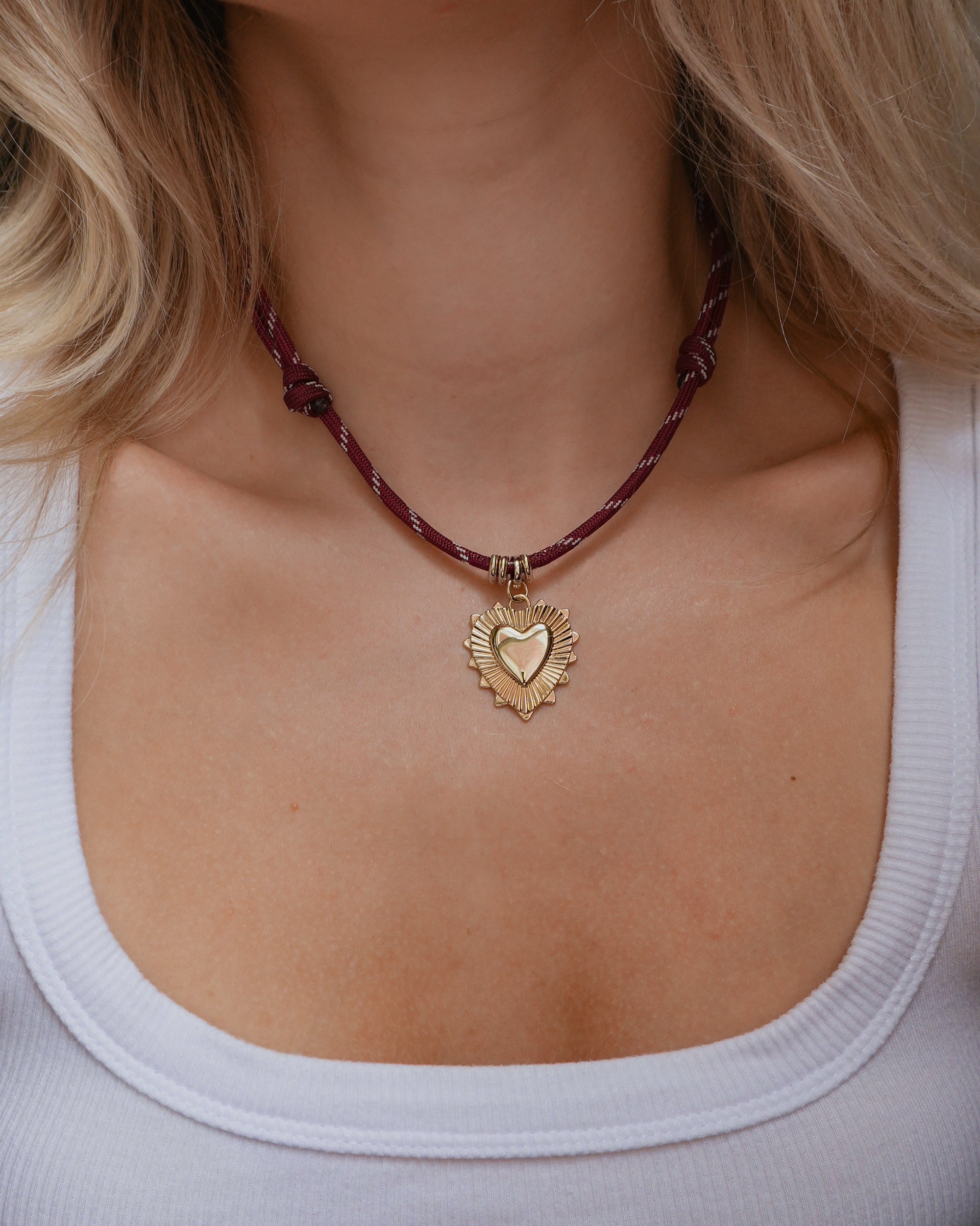 A woman wearing a white ribbed tank top and the Mod + Jo Tommy Cord Necklace—a maroon paracord necklace featuring gold plate accents and a gold heart-shaped pendant; only her neck, collarbone, and part of her hair are visible.