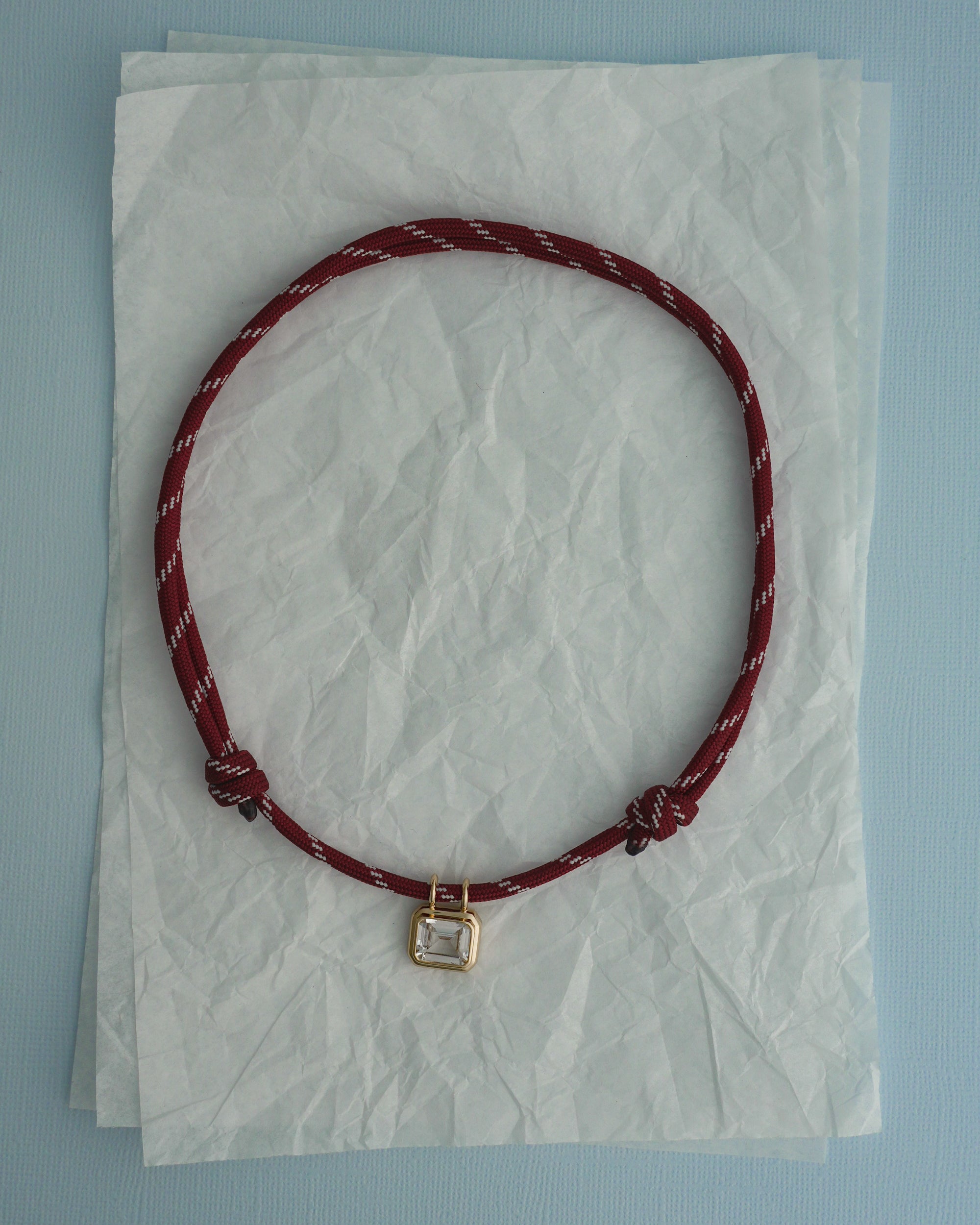 The Mod + Jo Tommy Sapphire Pendant Necklace, featuring a red adjustable paracord with white accents and a rectangular gold bezel pendant, is displayed on crumpled white tissue paper over a light blue background.