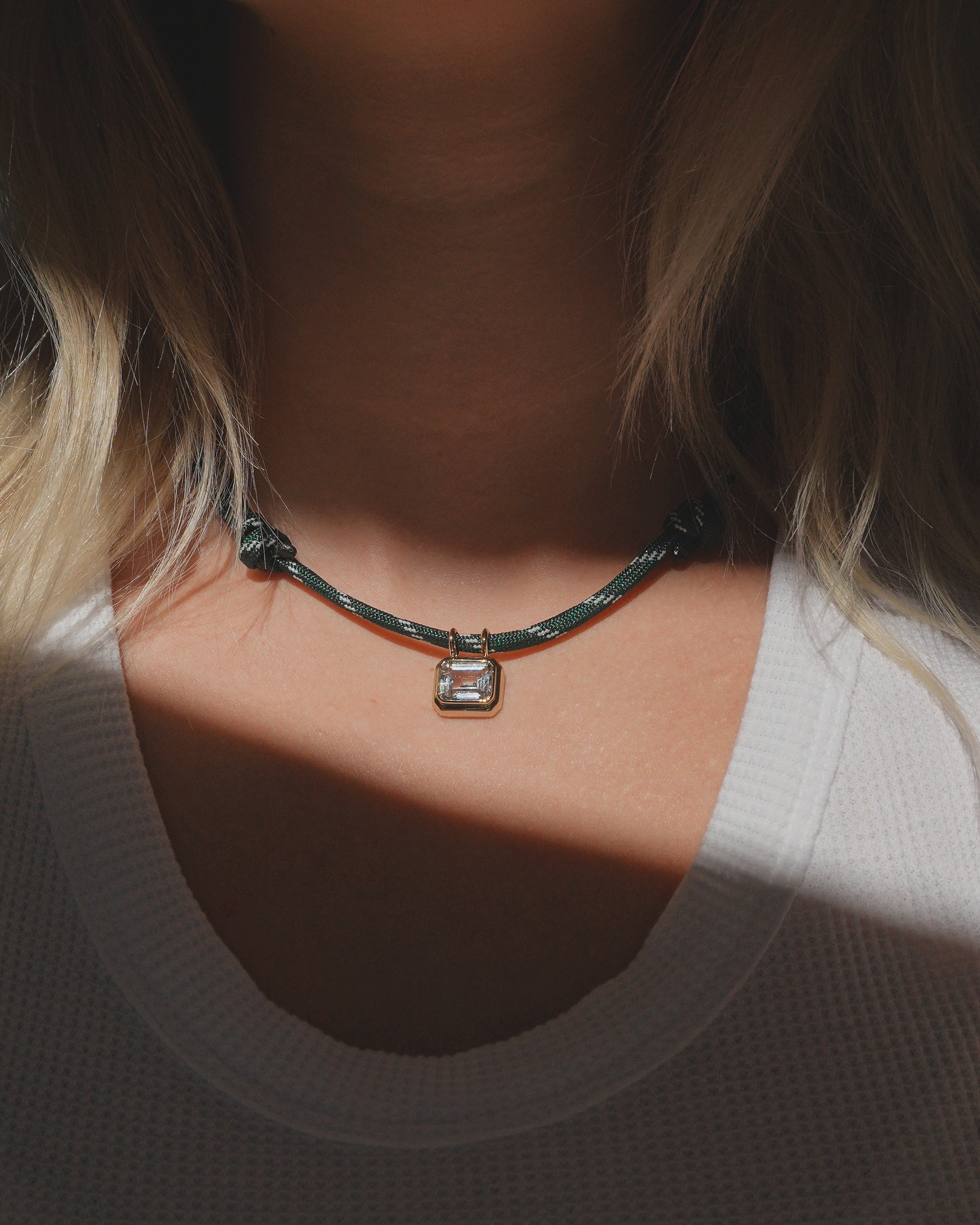 A person wearing the Mod + Jo Tommy Sapphire Pendant Necklace, featuring an adjustable green paracord and a silver padlock pendant, is shown from the neck down in a white textured shirt with sunlight casting shadows.