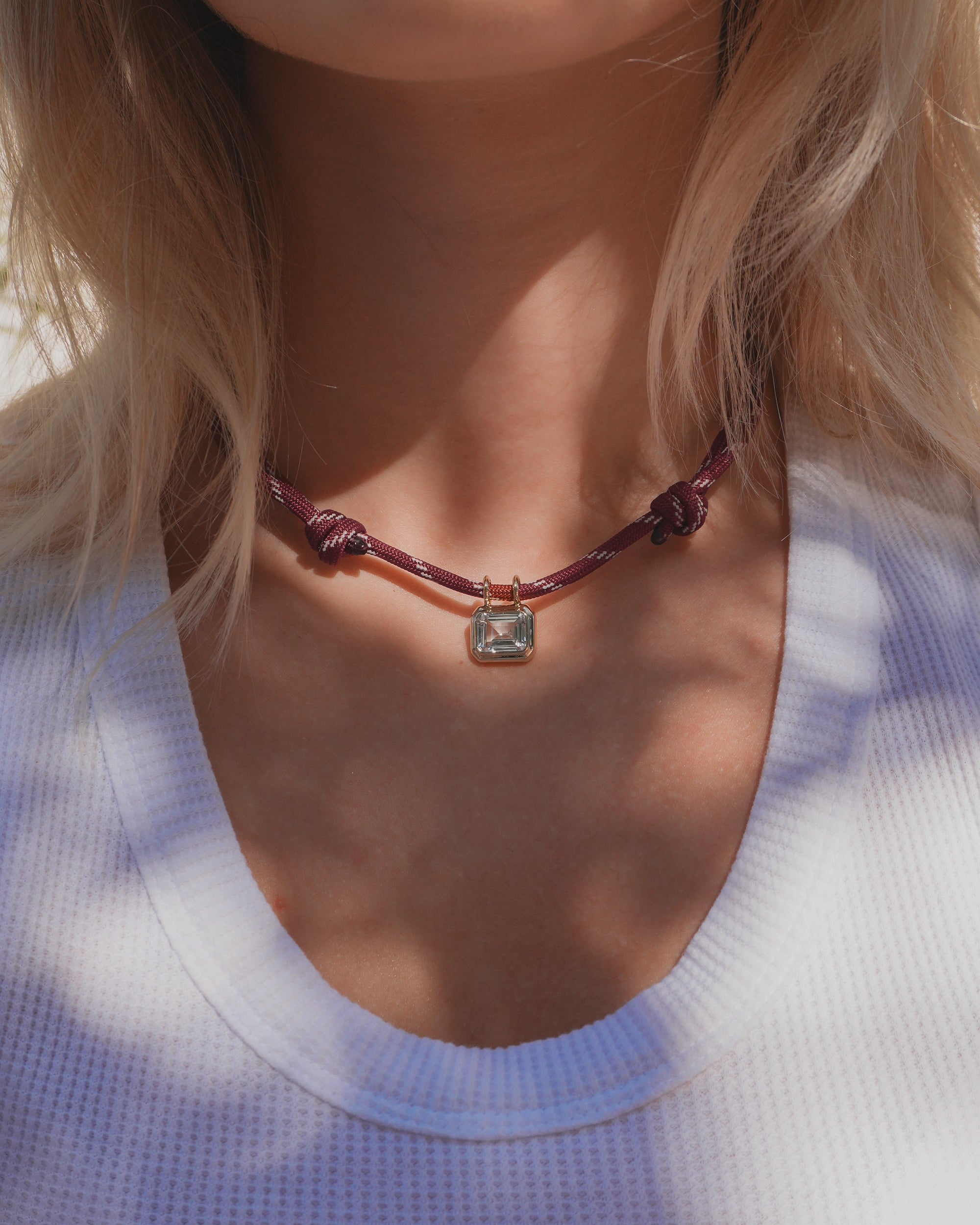 A person wearing a white textured shirt and the Mod + Jo Tommy Sapphire Pendant Necklace—a purple adjustable paracord necklace with a square lab-grown white sapphire pendant set in a gold bezel—shown from the shoulders to just below the chin.