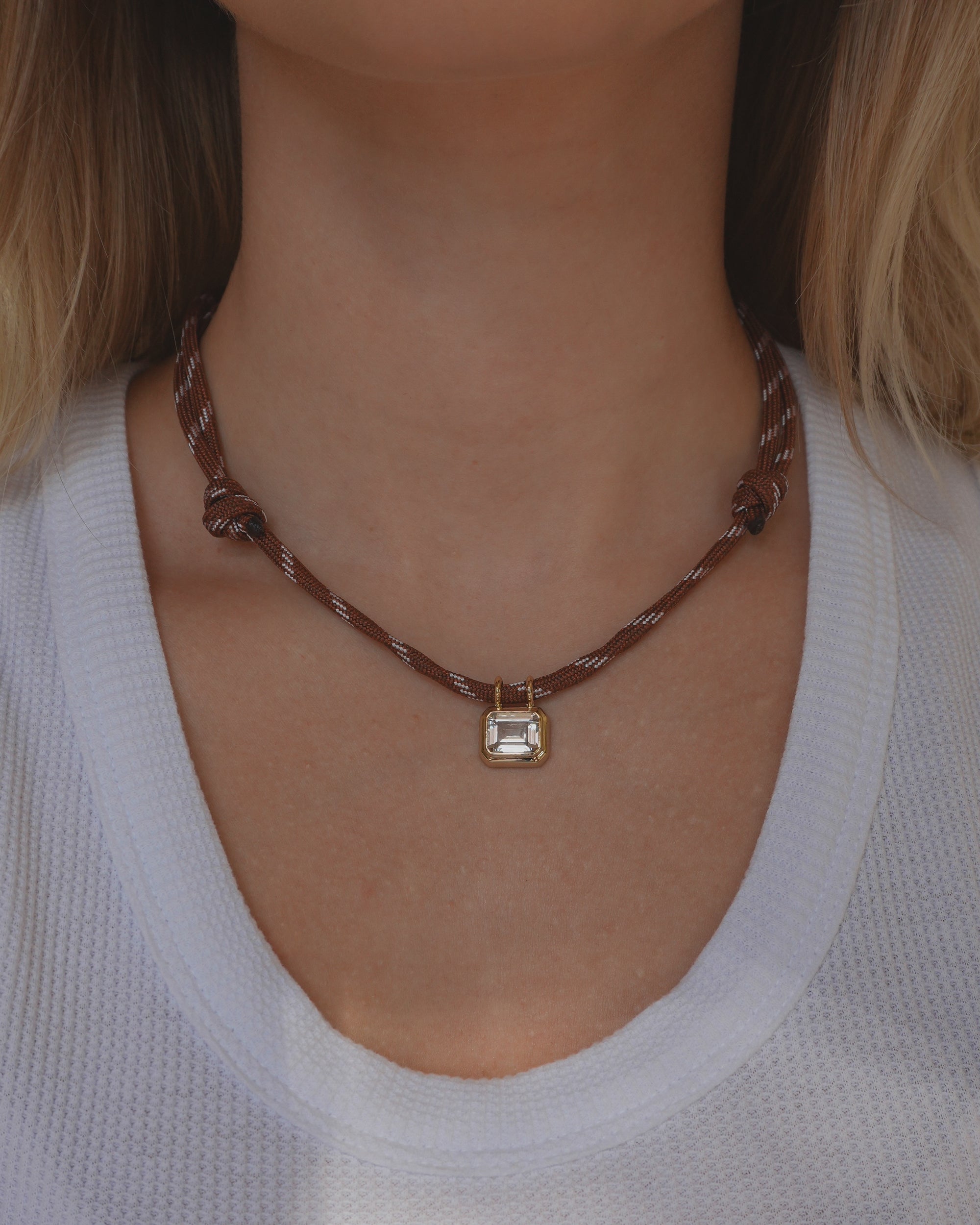 A person wearing the Mod + Jo Tommy Sapphire Pendant Necklace—an adjustable paracord necklace with a rectangular gold bezel-set pendant—paired with a white textured top.