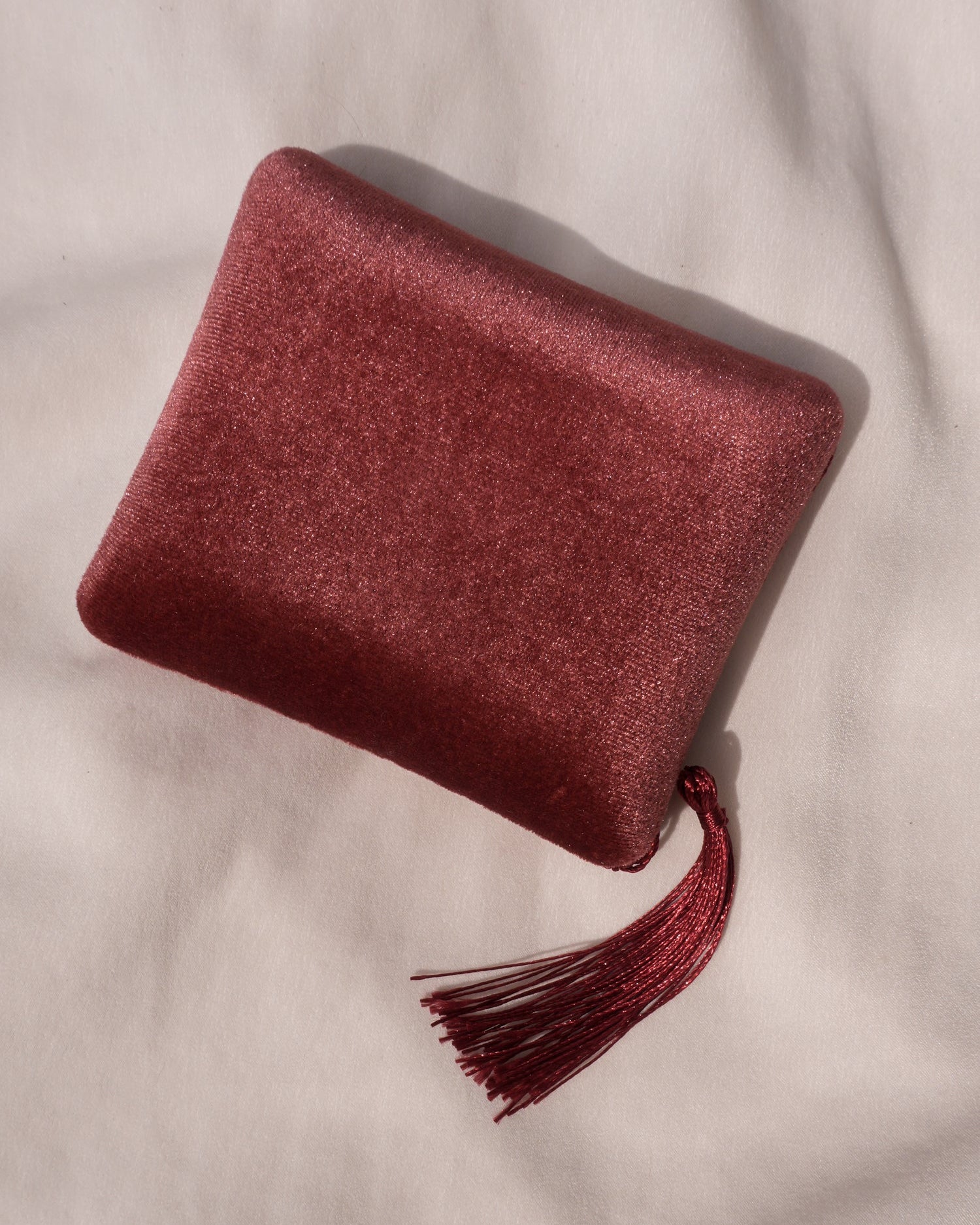 The Mod + Jo Tassel Velvet Jewelry Box, a small rectangular shimmery burgundy pouch with a matching tassel, sits atop a soft light fabric, its satin interior promising luxurious gift storage.