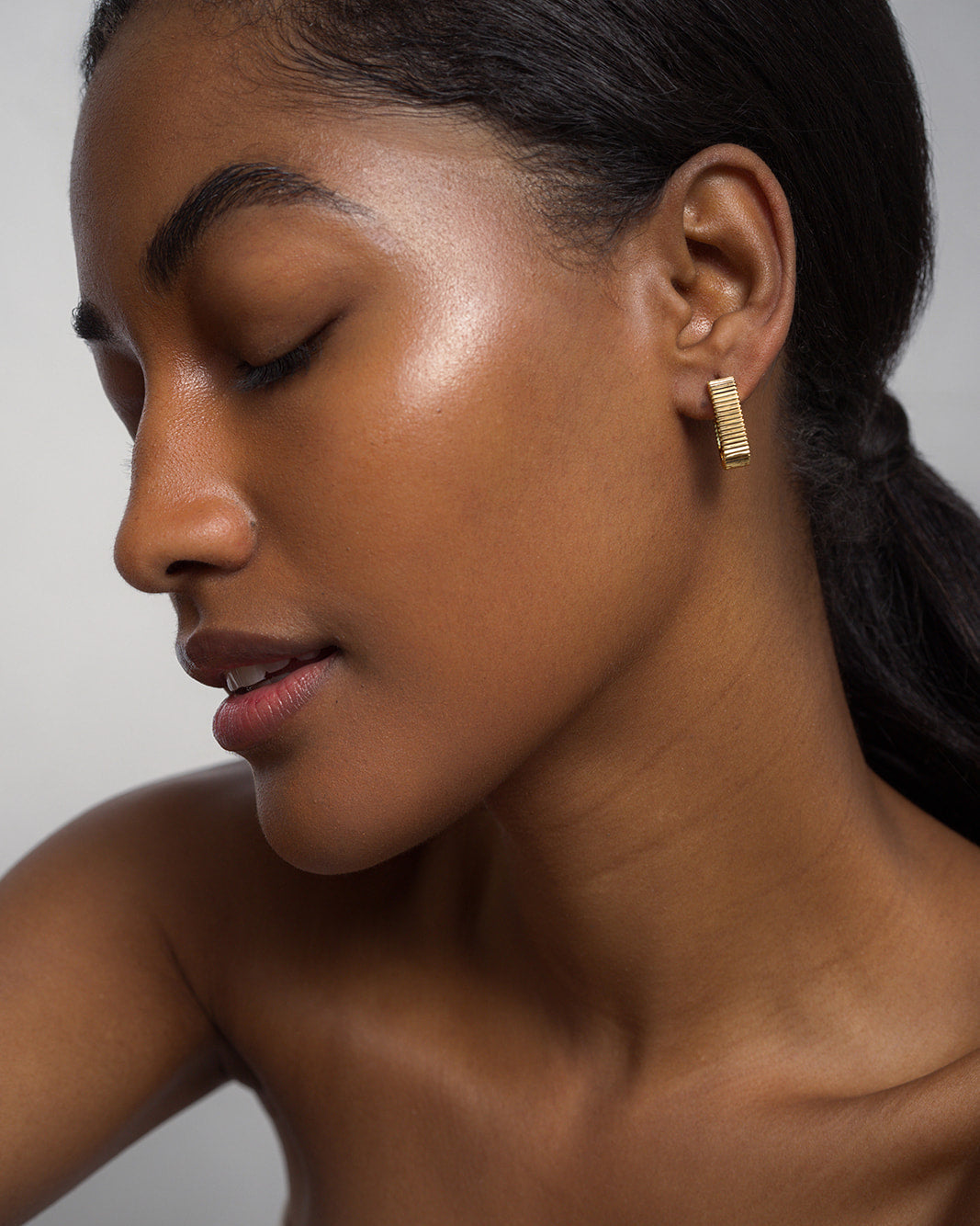 A woman with smooth, glowing skin and dark hair pulled back models the Elly Fluted Hoops by Mod + Jo. With her eyes closed and a gentle smile, she is photographed against a plain light background, highlighting the lightweight gold earrings.