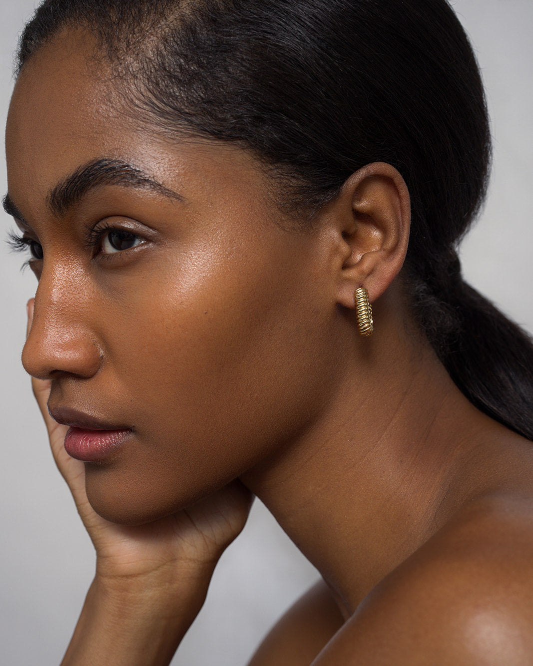 A woman with smooth, glowing skin rests her chin on her hand, gazing thoughtfully to the side. She wears Mod + Jo’s Elly French Rope Hoops that catch the light, and her eyebrows are neatly groomed.