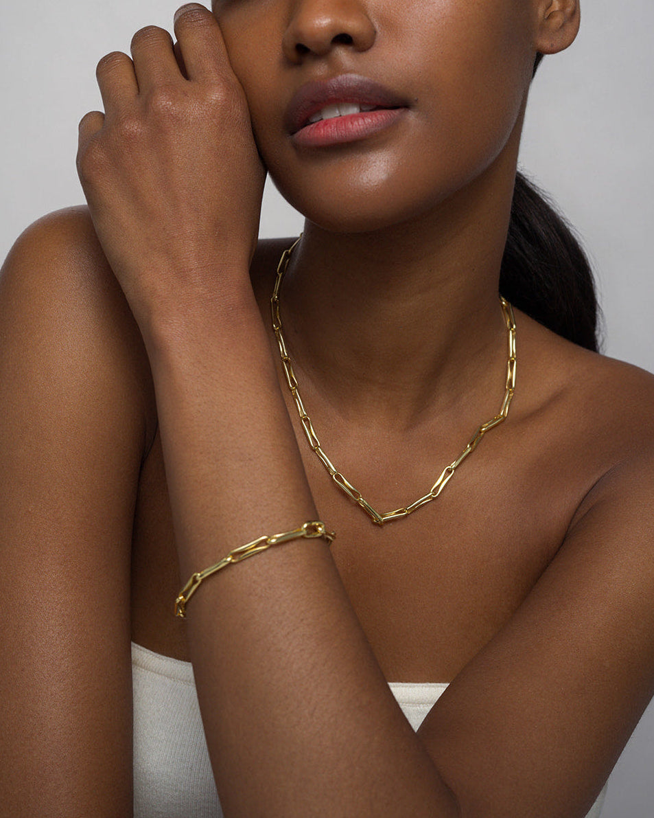 A close-up of a model wearing a gold chain necklace and a matching gold bracelet against a neutral gray background.