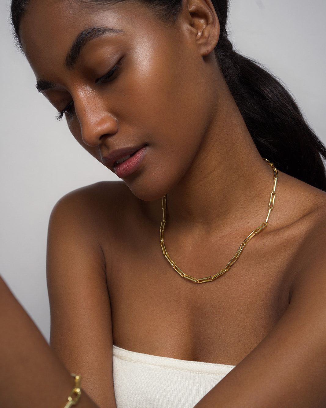 A woman with smooth dark skin and long dark hair wears the Mod + Jo Molly Elongated Link Chain Necklace along with a matching bracelet, looking down with a calm expression. She is dressed in a white strapless top against a plain background.