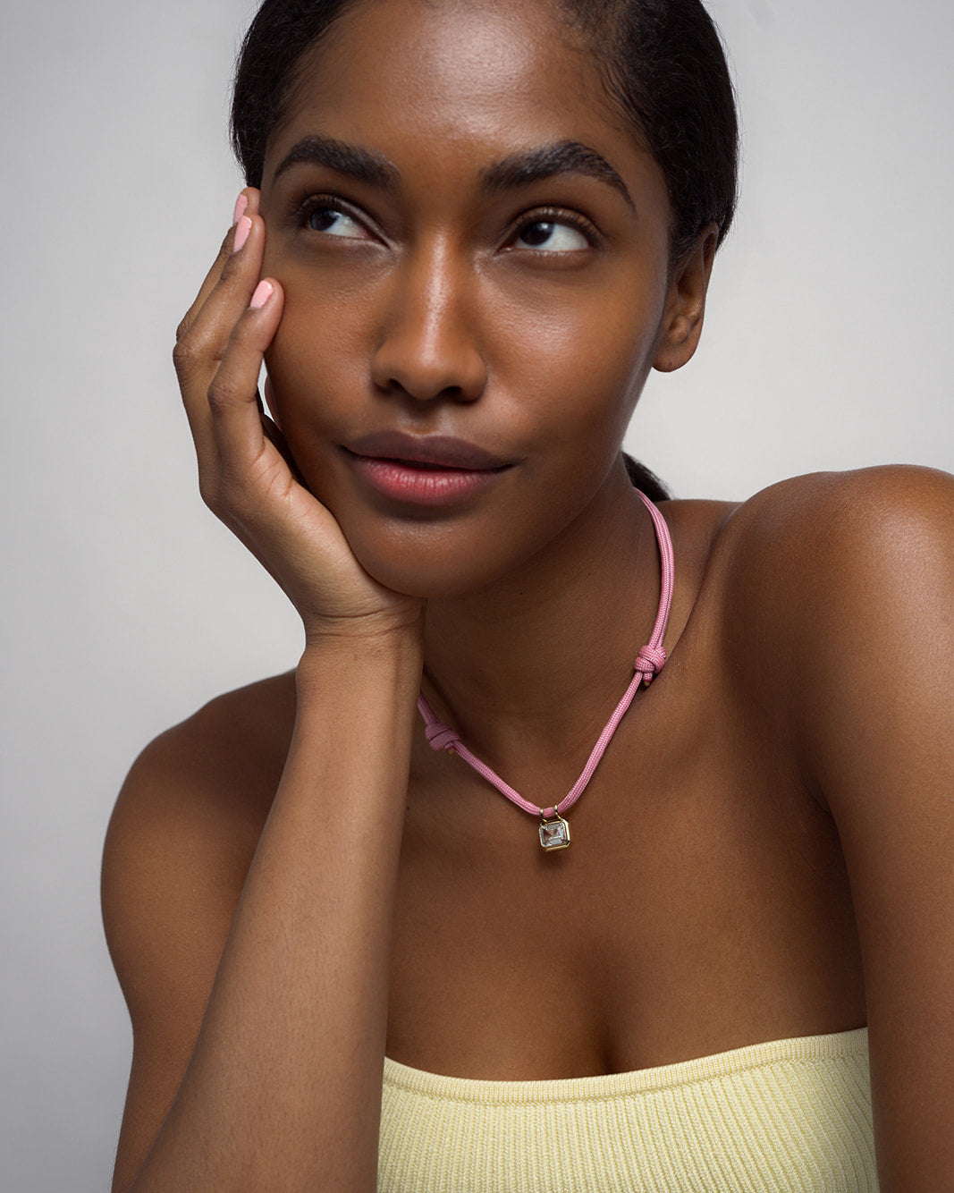 A woman with smooth brown skin and straight dark hair rests her face on her hand, wearing a pale yellow top and the Mod + Jo Tommy Sapphire Pendant Necklace—a pink adjustable paracord necklace featuring a small pendant—while gazing thoughtfully upward against a plain background.