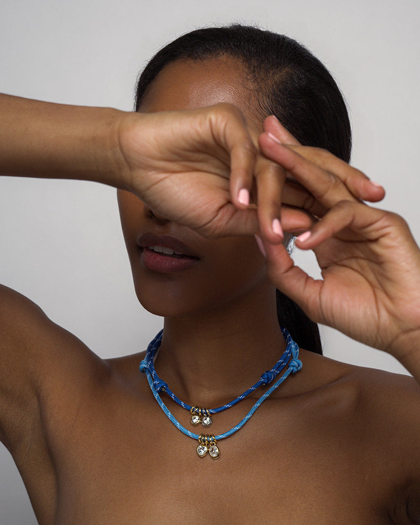 A woman wearing the Mod + Jo Tommy Sapphire Pendant Necklace, featuring layered blue adjustable paracord with jeweled accents and a gold bezel pendant, poses with her arm partially covering her face against a plain background.