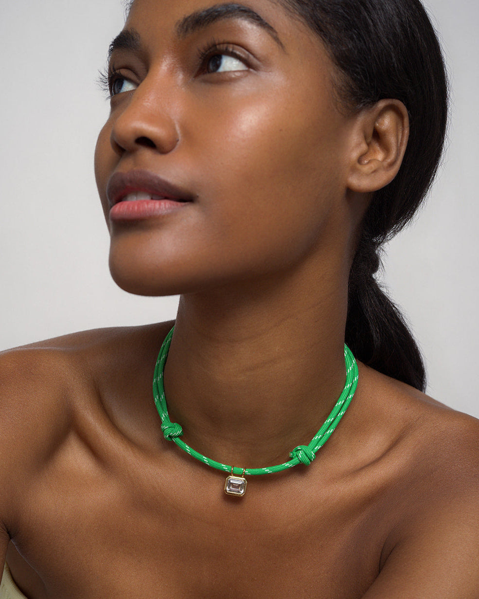A woman looks upward, wearing the Mod + Jo Tommy Sapphire Pendant Necklace—a green adjustable paracord necklace with a small metallic pendant. She has smooth skin, natural makeup, and dark hair pulled back, set against a plain light background.