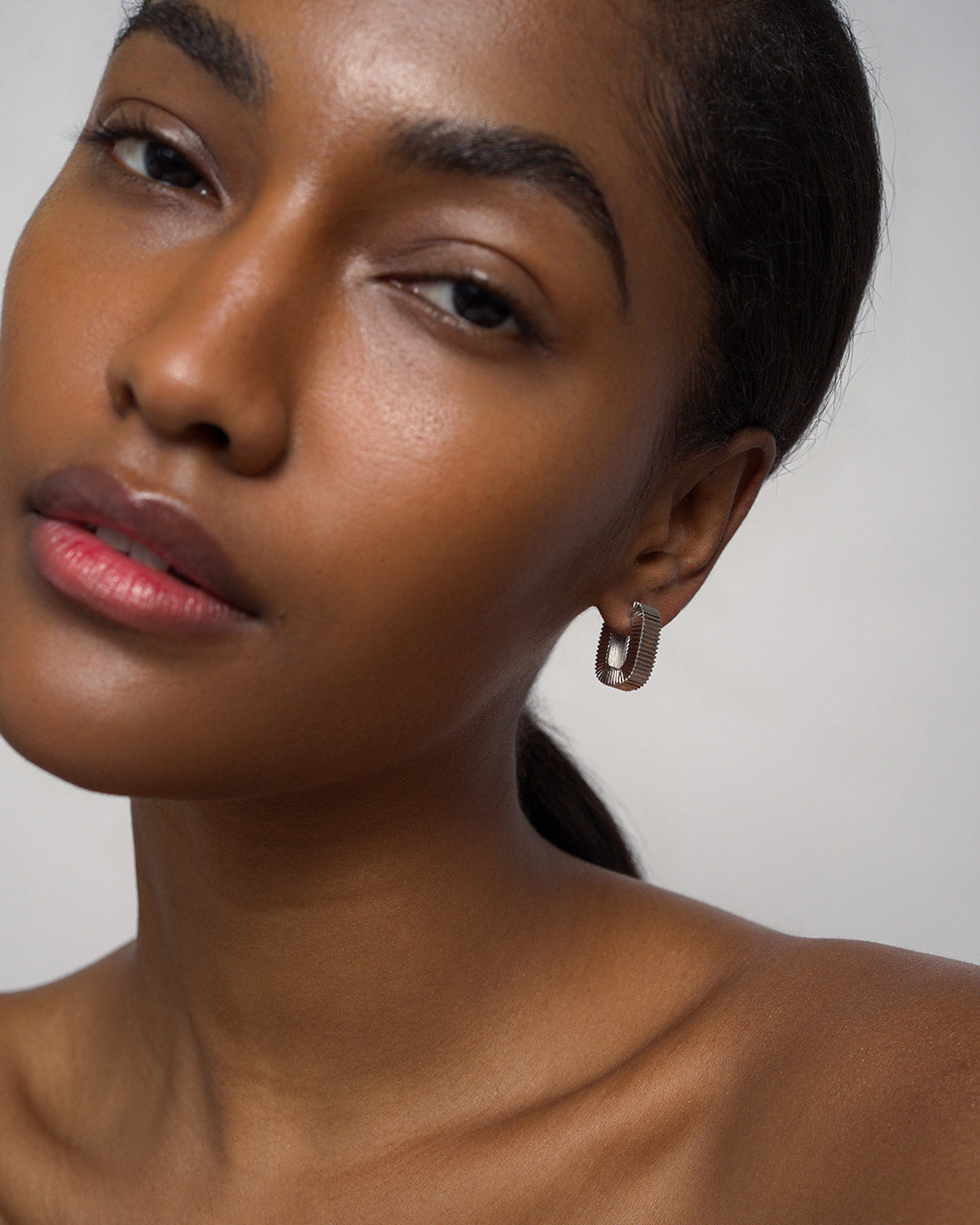 A woman with smooth brown skin and dark hair pulled back gazes softly at the camera. She wears a single, chunky Elly Fluted Hoop by Mod + Jo, paired with natural makeup and bare shoulders against a plain background. The bold silver earring adds a touch of modern elegance to her understated look.