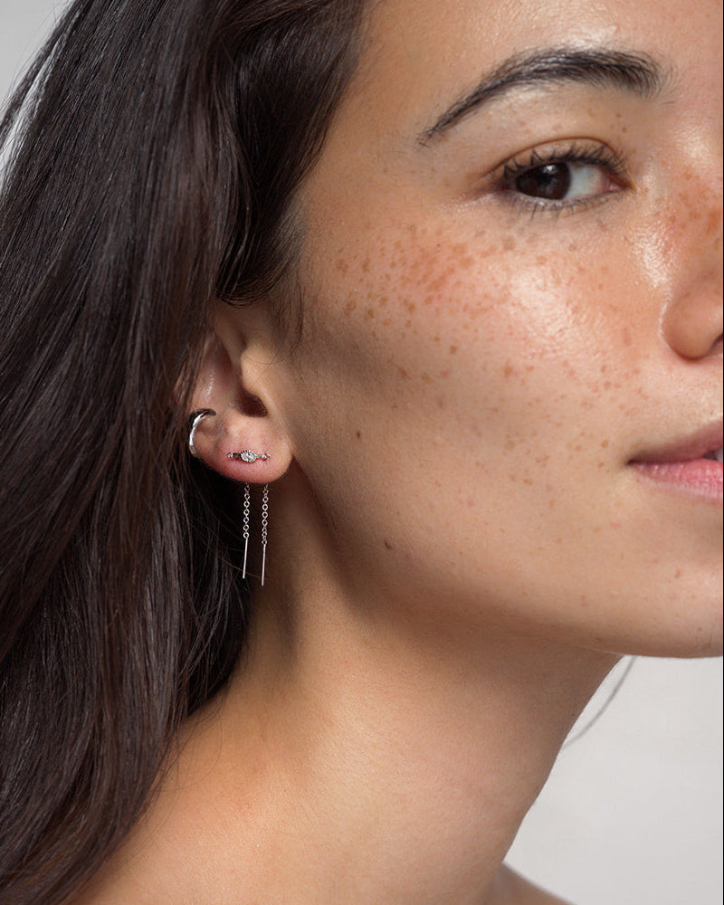 Close-up of a woman’s face, highlighting her ear adorned with three earrings: a small rhodium-plated hoop, a sparkling cubic zirconia stud, and two long Nicole Oval Threader Earrings by Mod + Jo. She has smooth, freckled skin and dark hair tucked behind her ear.