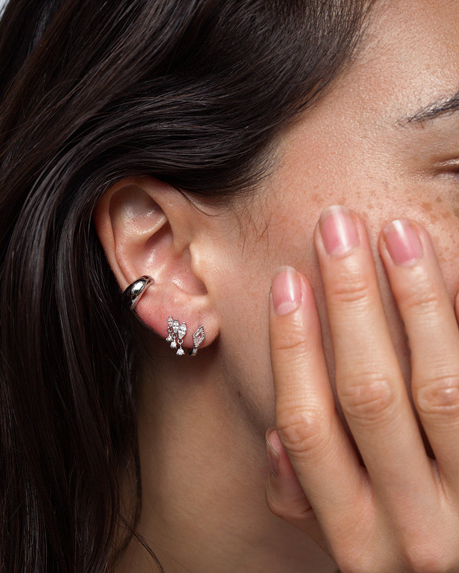 A close-up of a person’s ear adorned with three earrings: a silver hoop at the top and two gemstone studs in the lobe, one of which is Mod + Jo’s Gabby Marquise Dangle Studs that catch the light beautifully. The person’s neatly manicured hand rests gently against their face.