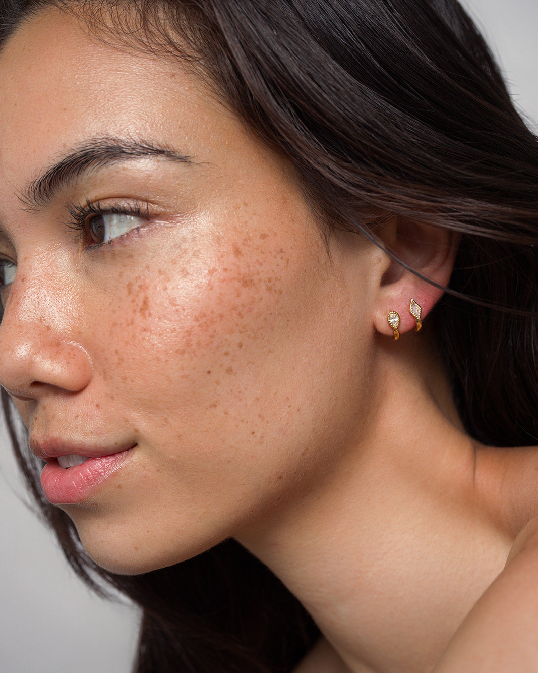 A close-up of a woman’s face shows her natural freckles and smooth skin as she looks off to the side against a plain background. She wears Mod + Jo’s Phoebe Kite Huggies—small, gold-plated, leaf-shaped earrings accented with sparkling cubic zirconia.