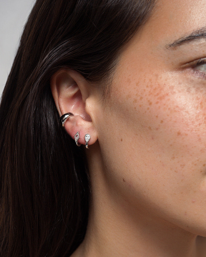 A close-up of a woman’s ear with three earrings: a small rhodium plated hoop on the upper ear, and two huggie earrings with gem-studded hoops in the lobe—one being the Mod + Jo Phoebe Kite Huggies, featuring a delicate kite-cut cubic zirconia. She has straight, dark hair and light skin with freckles.
