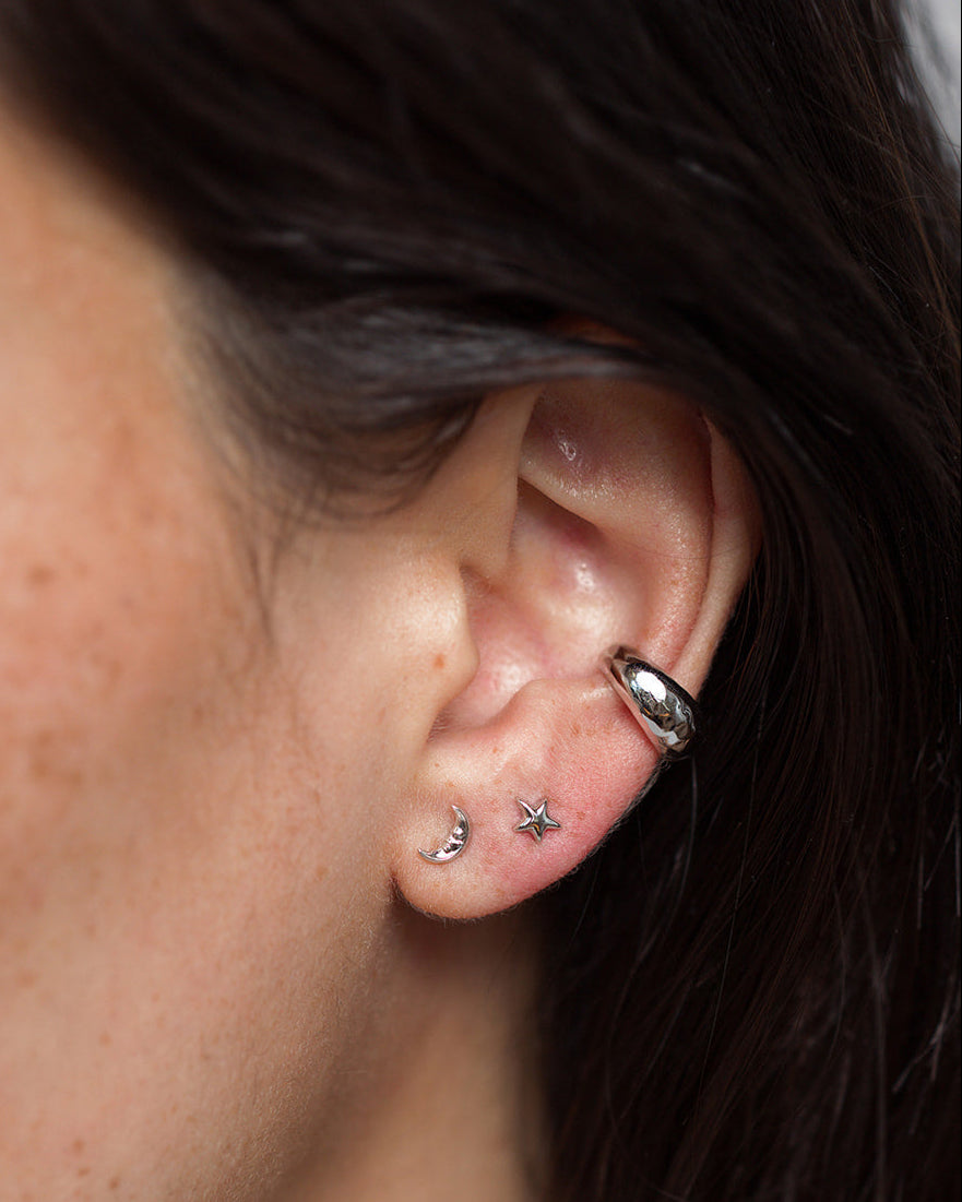 Close-up of an ear with three piercings: a sleek rhodium-plated silver hoop on the upper ear, and two celestial earrings—the Mod + Jo Archie Moon Studs, shaped like a crescent moon and a star—adorning the lower lobe. Dark hair partially frames the ear.