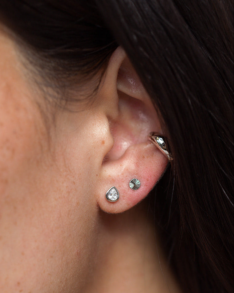Close-up of a person’s ear adorned with three earrings: a small rhodium-plated stud, the Mod + Jo Joni Bezel Studs featuring a circular textured bezel design, and a silver hoop cuff on the upper ear. The person’s dark hair partially covers the ear.