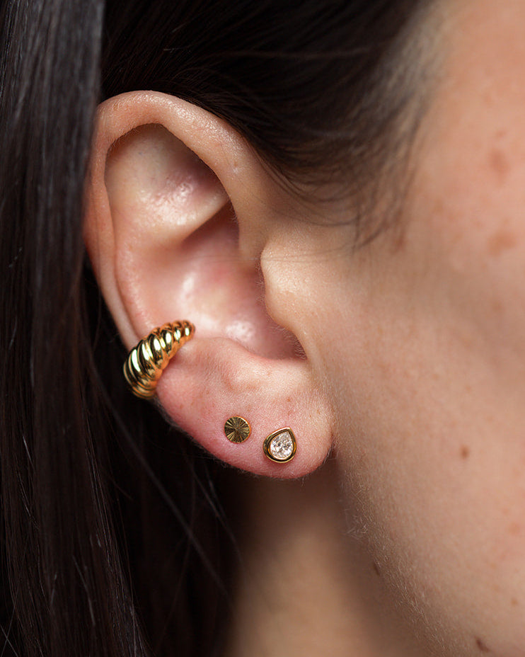 A close-up of a person’s ear adorned with three gold plated earrings: a spiral hoop on the upper ear, a small sunburst stud, and the Joni Bezel Stud by Mod + Jo featuring a clear cubic zirconia stone on the lobe. The person has straight, dark hair.
