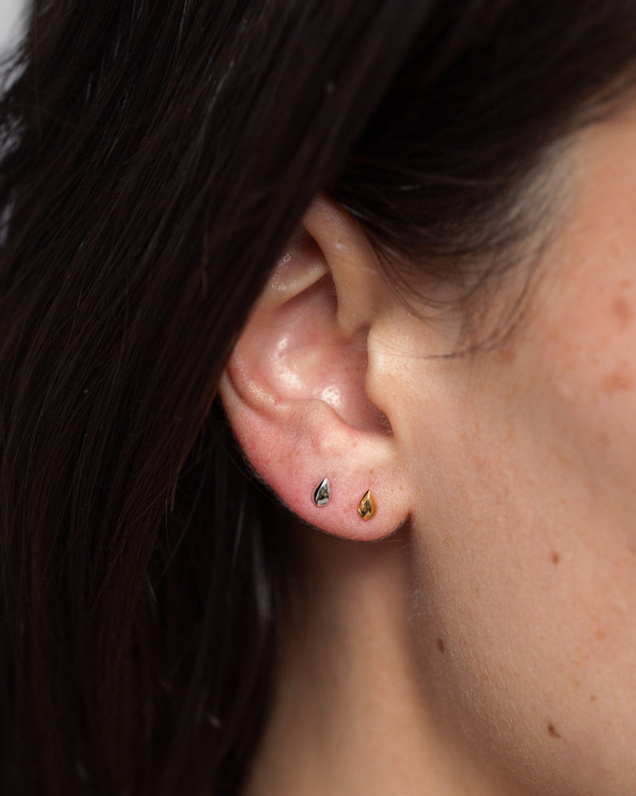 A close-up of a person’s ear features the Uma Teardrop Studs by Mod + Jo—one in gold and one in silver—worn in the earlobe. With dark hair tucked behind the ear, these elegant teardrop-shaped earrings are perfectly showcased, making them an effortless choice for everyday jewelry.
