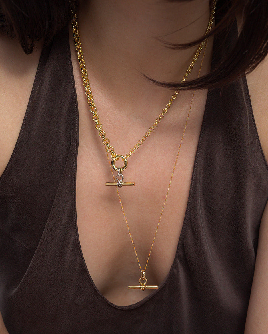 A person wearing a dark, deep V-neck top showcases two layered Mod + Jo Elliot Toggle Charm necklaces in 14k gold, each featuring a horizontal bar pendant and distinct chain styles, set against their bare upper chest.