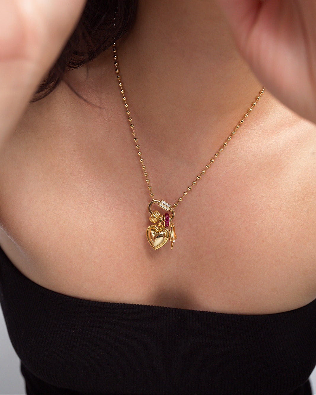 A woman wearing the Mod + Jo Nora Lock Carabiner necklace, featuring a gold 14k-plated delicate chain adorned with a heart-shaped pendant accented by a petite blue lace agate gemstone and an additional tiny charm, is shown close up against her bare collarbone and a black off-shoulder top. The semi-precious stones and gleaming finish highlight the necklace’s elegant design.