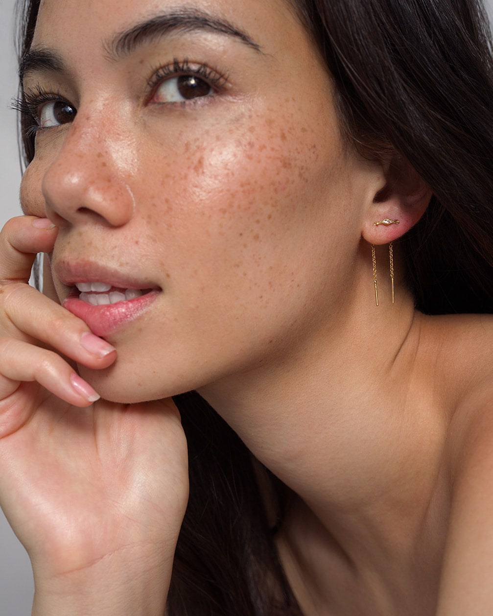 A woman with long dark hair and freckled skin poses confidently with her hand near her chin, wearing the Mod + Jo Nicole Kite Threader Earring in delicate gold plating.
