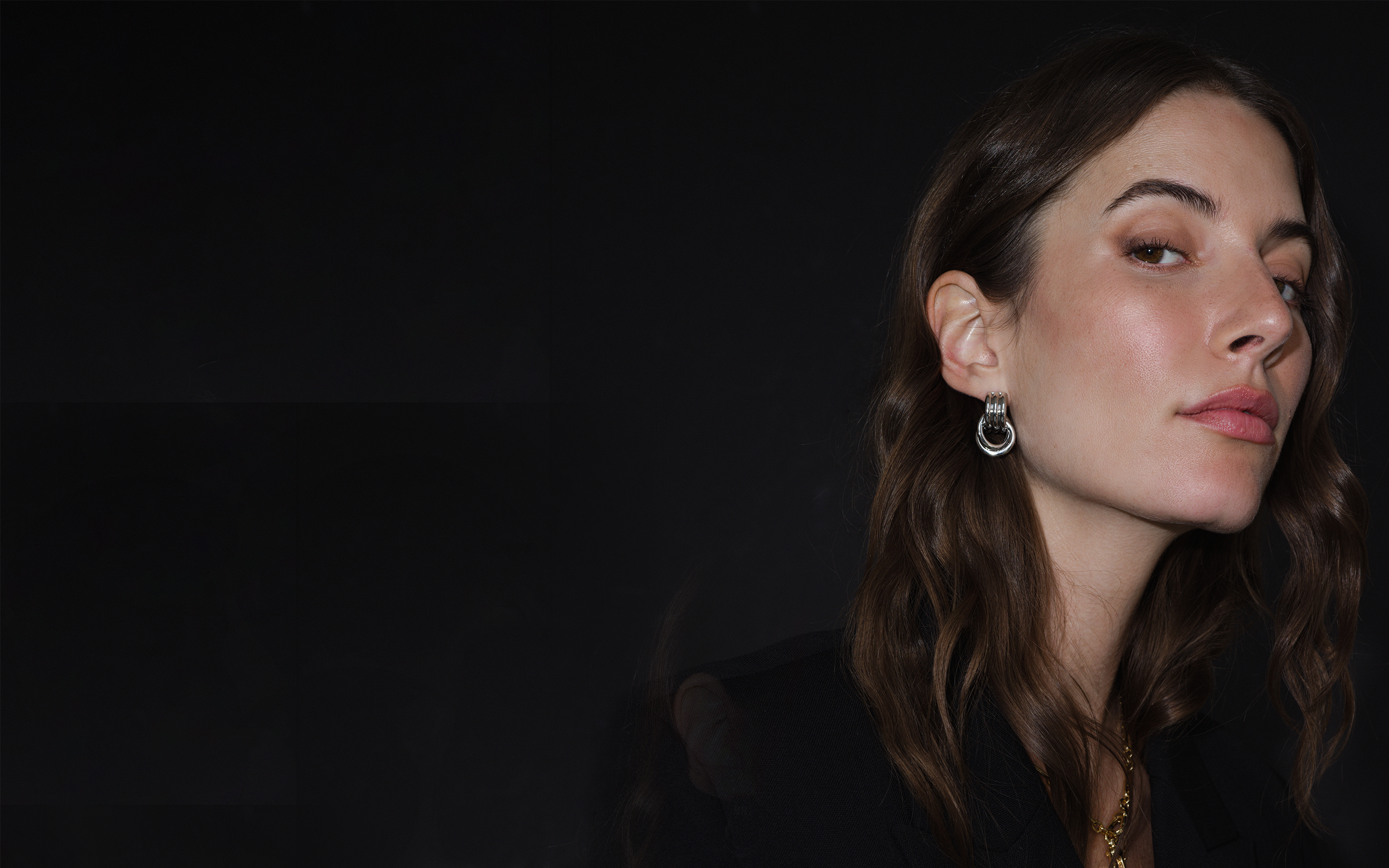 Woman with long brown hair wearing earrings against a dark background