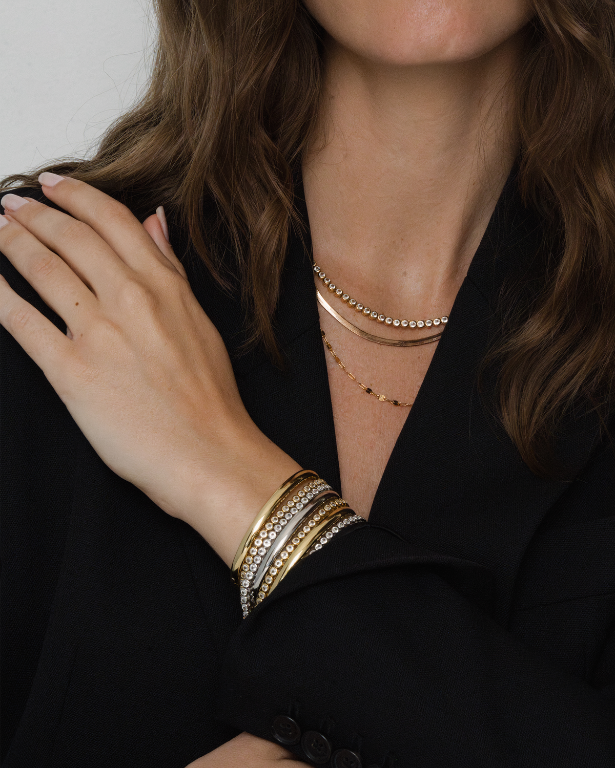 The Emma Set by Mod + Jo, featuring three layered gold necklaces including a bezel tennis necklace and a stack of gold and rhinestone tennis bracelets, worn with a black blazer.