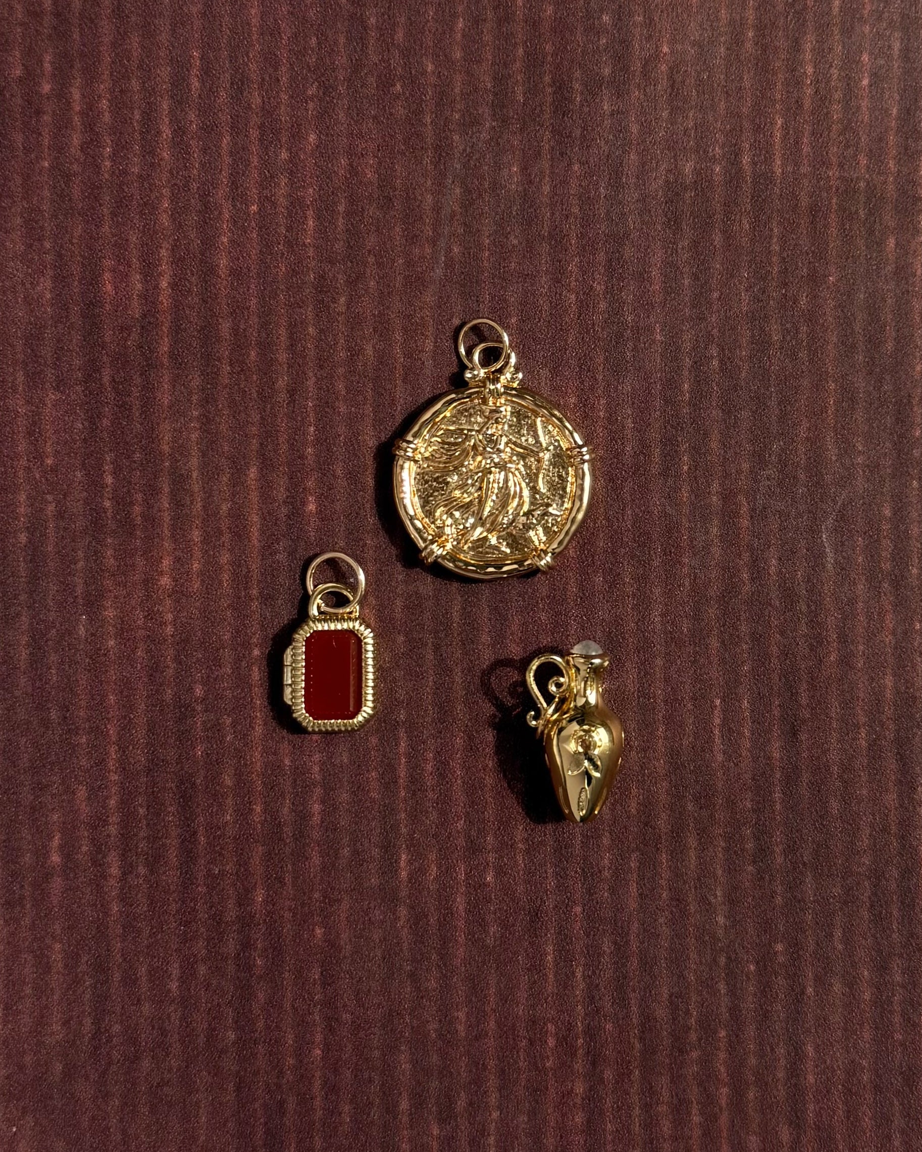 Three 14k gold-plated charms from Mod + Jo rest on a dark, textured surface: a round charm with an intricate design, the Mabel Rectangle Birthstone Locket Charm with a red center, and a heart-shaped charm featuring a small loop.