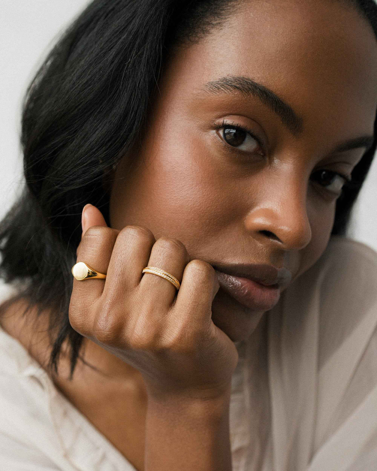 A woman rests her cheek against her hand, showcasing the Mariah + Tessa Set featuring a smooth gold signet ring and a thin pavé band with small embedded stones.