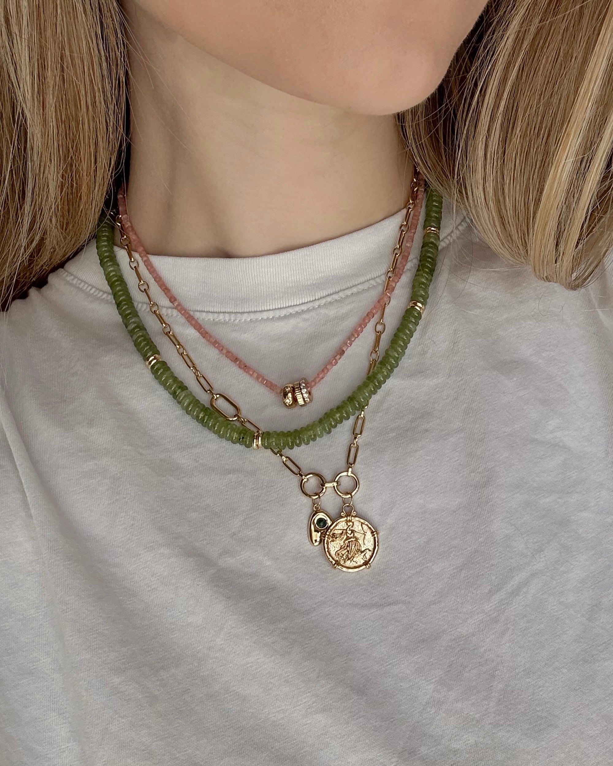 A person wears a white t-shirt with layered necklaces: the Mod + Jo Merit Gemstone Necklace in pink, a green beaded chain, and two 14k gold-filled chains—one with a coin pendant and one with a small lock charm.