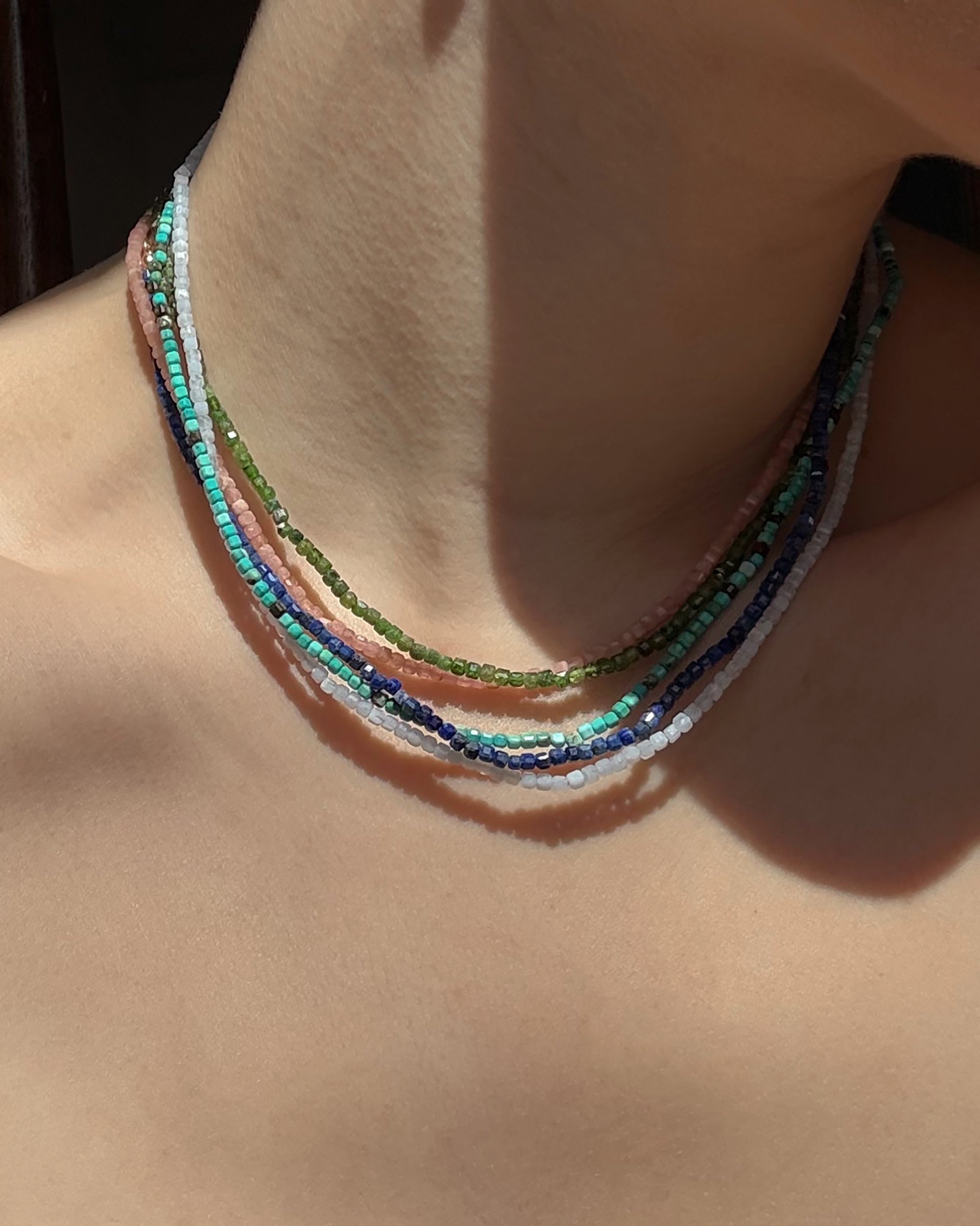 A close-up of a person’s neck shows several layered Mod + Jo Merit Gemstone Necklaces in hand-crafted white, green, blue, pink, and turquoise gemstones glowing against sunlit skin.