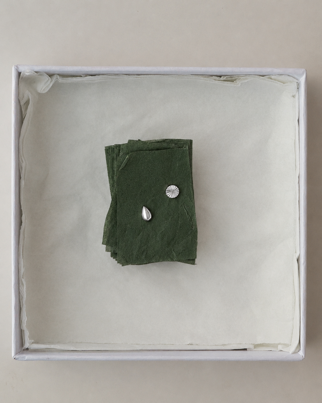 Dela + Uma Set featuring two mismatched silver stud earrings, one teardrop-shaped and one fluted sunburst-patterned, resting on a small stack of dark green paper inside a white box.
