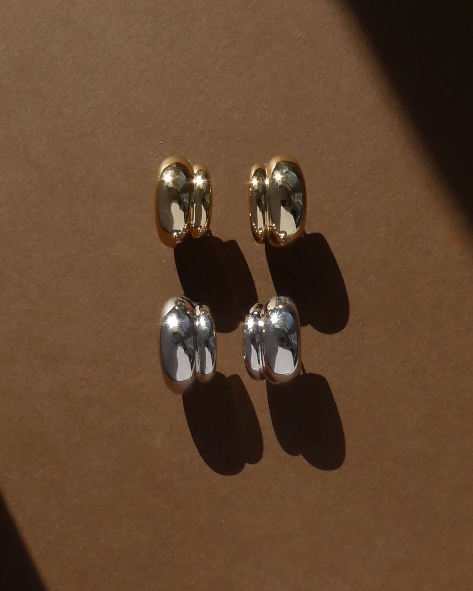 Four metallic cufflinks, two gold and two silver, are paired on a brown surface with soft shadows diagonally cast—a stylish arrangement that pairs perfectly with Mod + Jo’s Grande Sofie Volume Hoops.