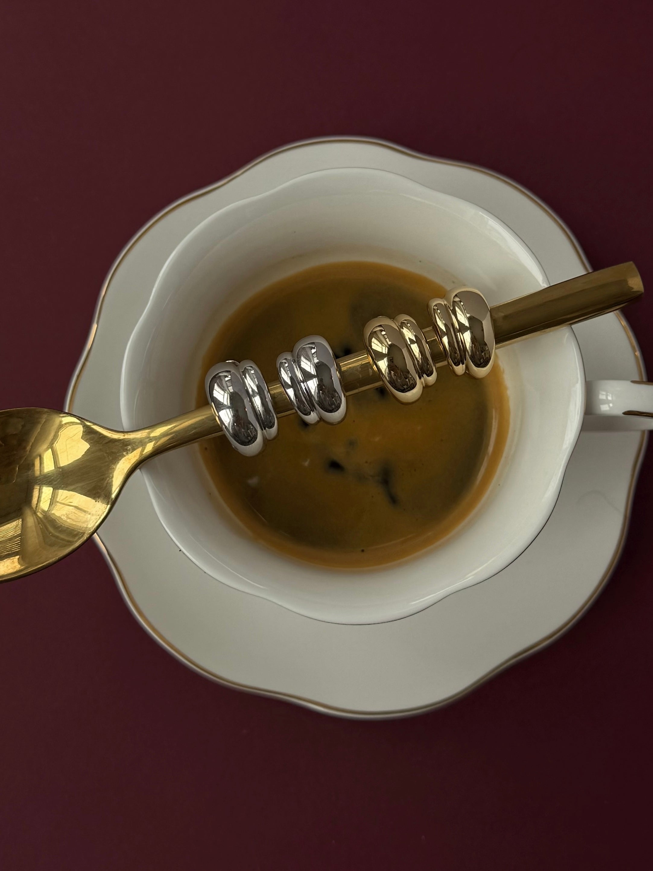 A cup of coffee on a white saucer with a gold spoon displays five Mod + Jo Petite Sofie Volume Hoops, their shiny metallic finish stacked elegantly on the spoon, set against a dark burgundy background.