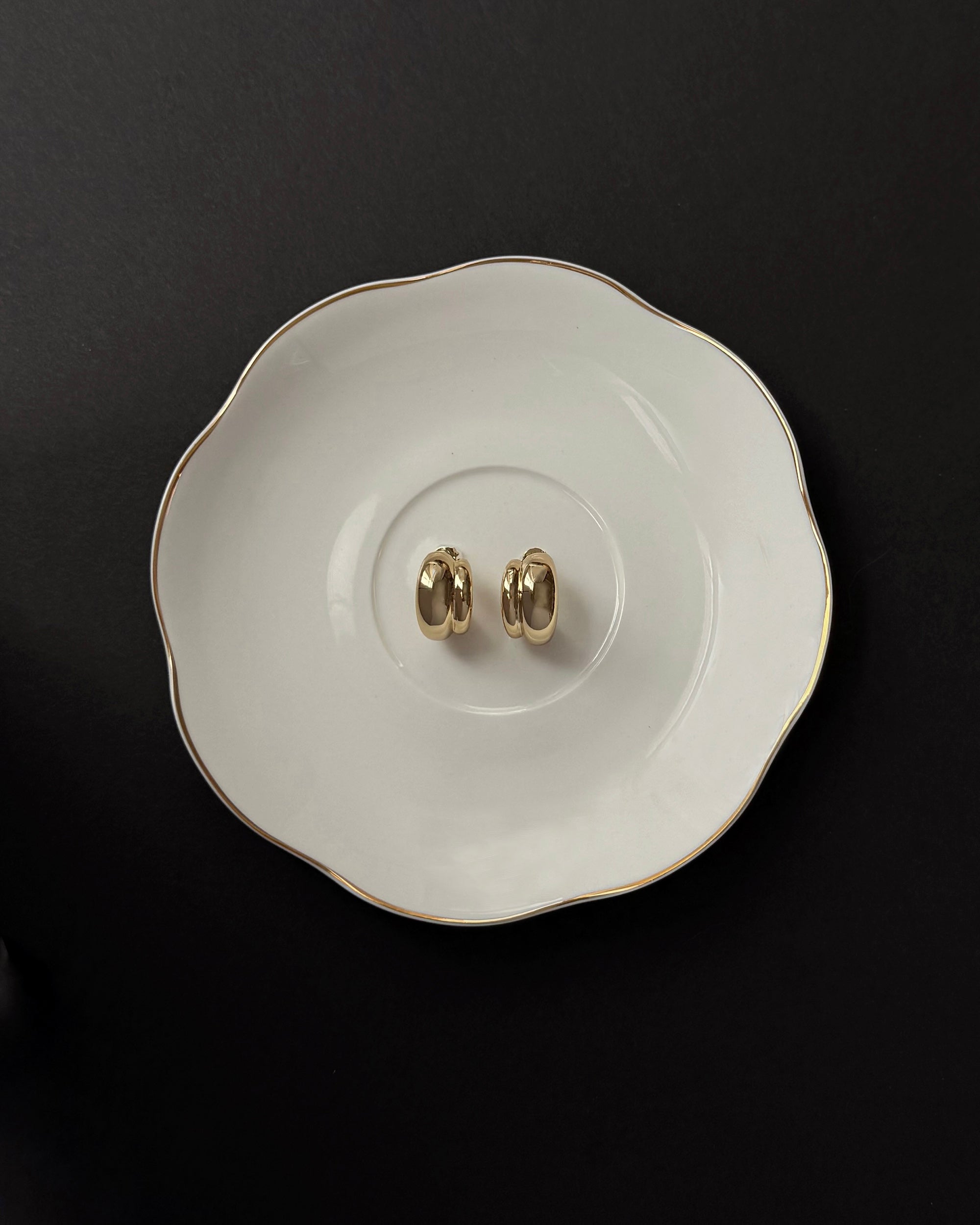 A white teacup tipped on its side spills out gold and silver hoop earrings of various sizes onto a black surface.