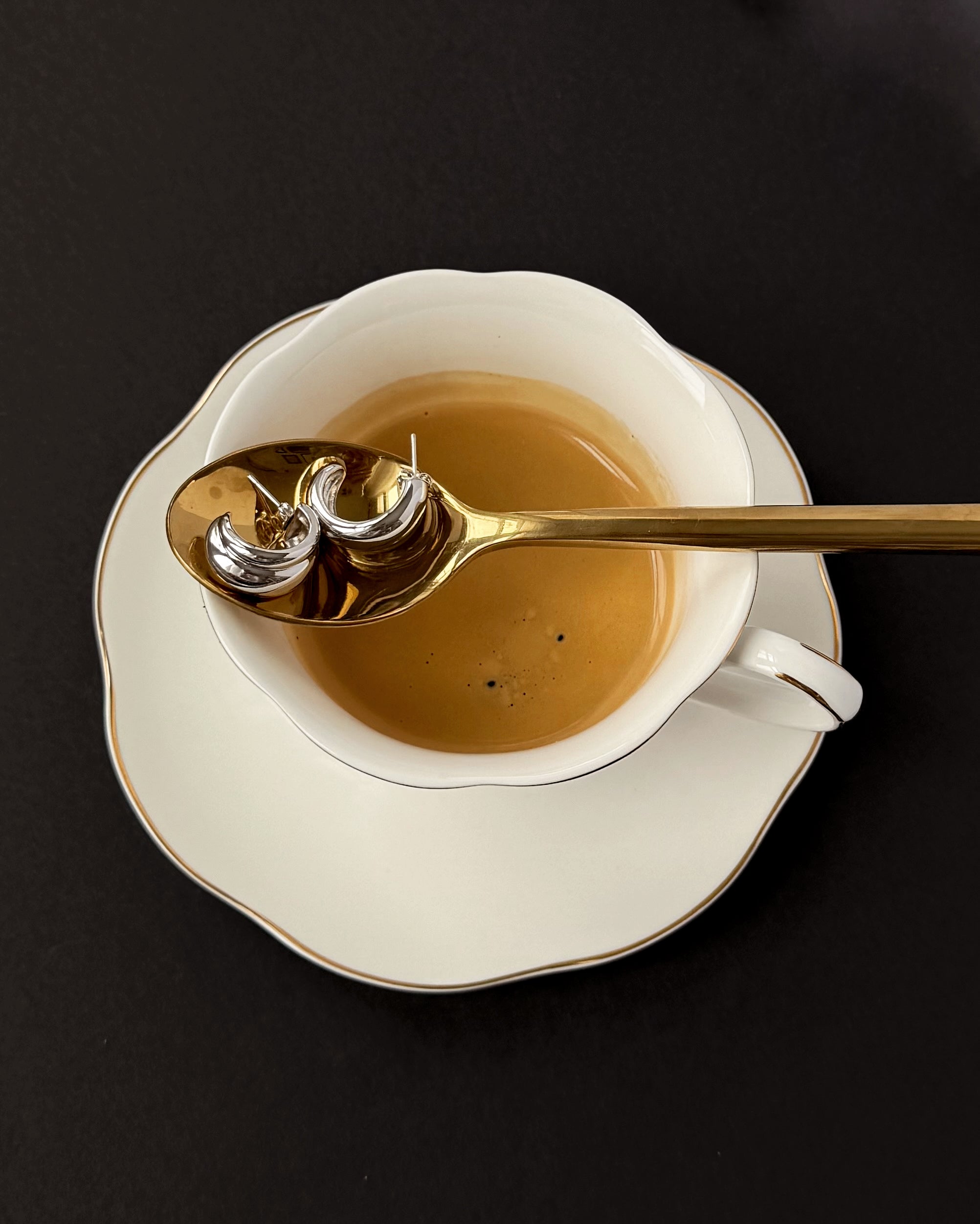 A gold spoon holding two silver rings rests over a white cup filled with coffee, placed on a matching saucer against a dark background.