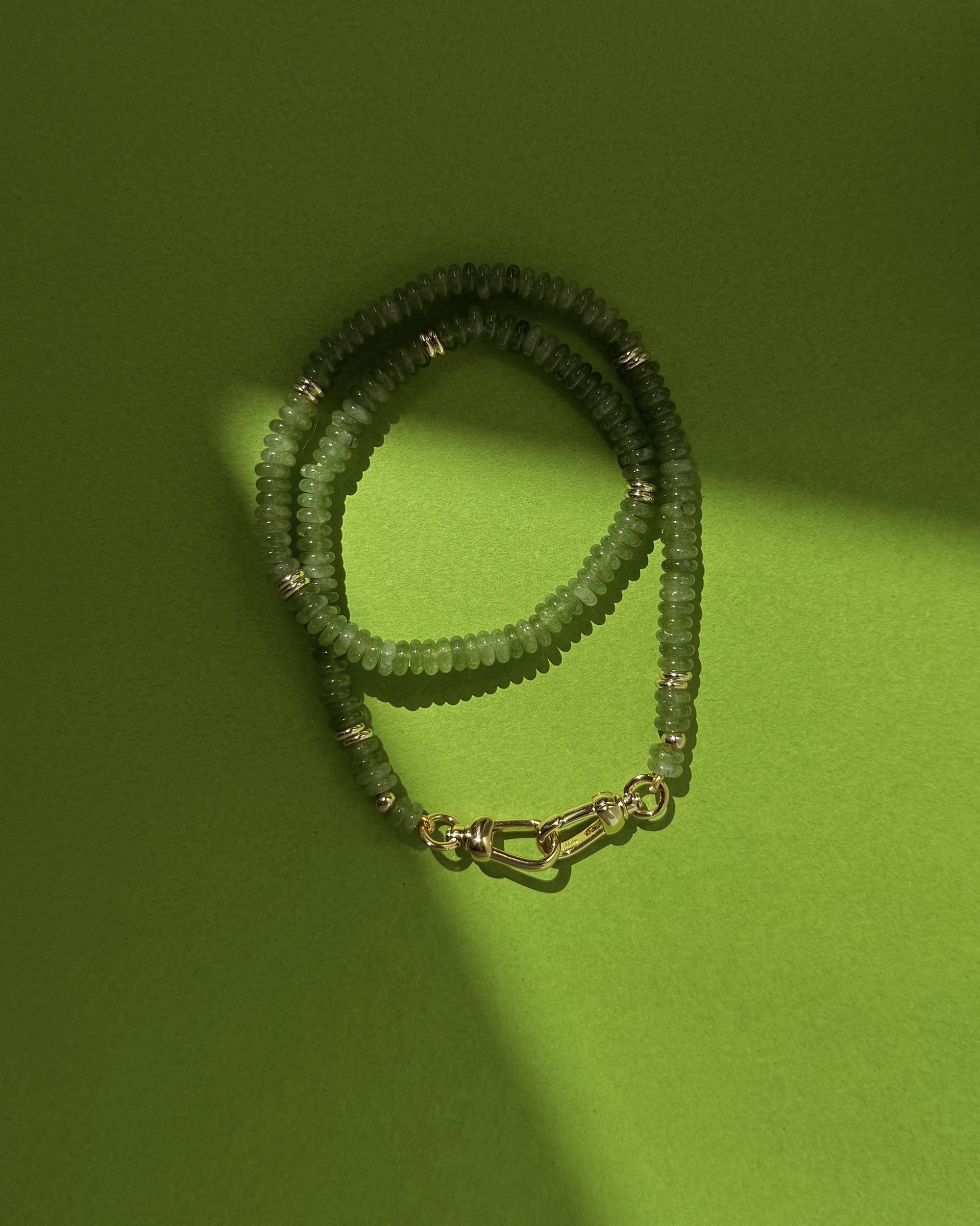 The Sonia Gemstone Necklace by Mod + Jo, featuring genuine gemstone beads, silver accents, and a silver clasp, is coiled on a bright green background with a diagonal shadow across part of the image.