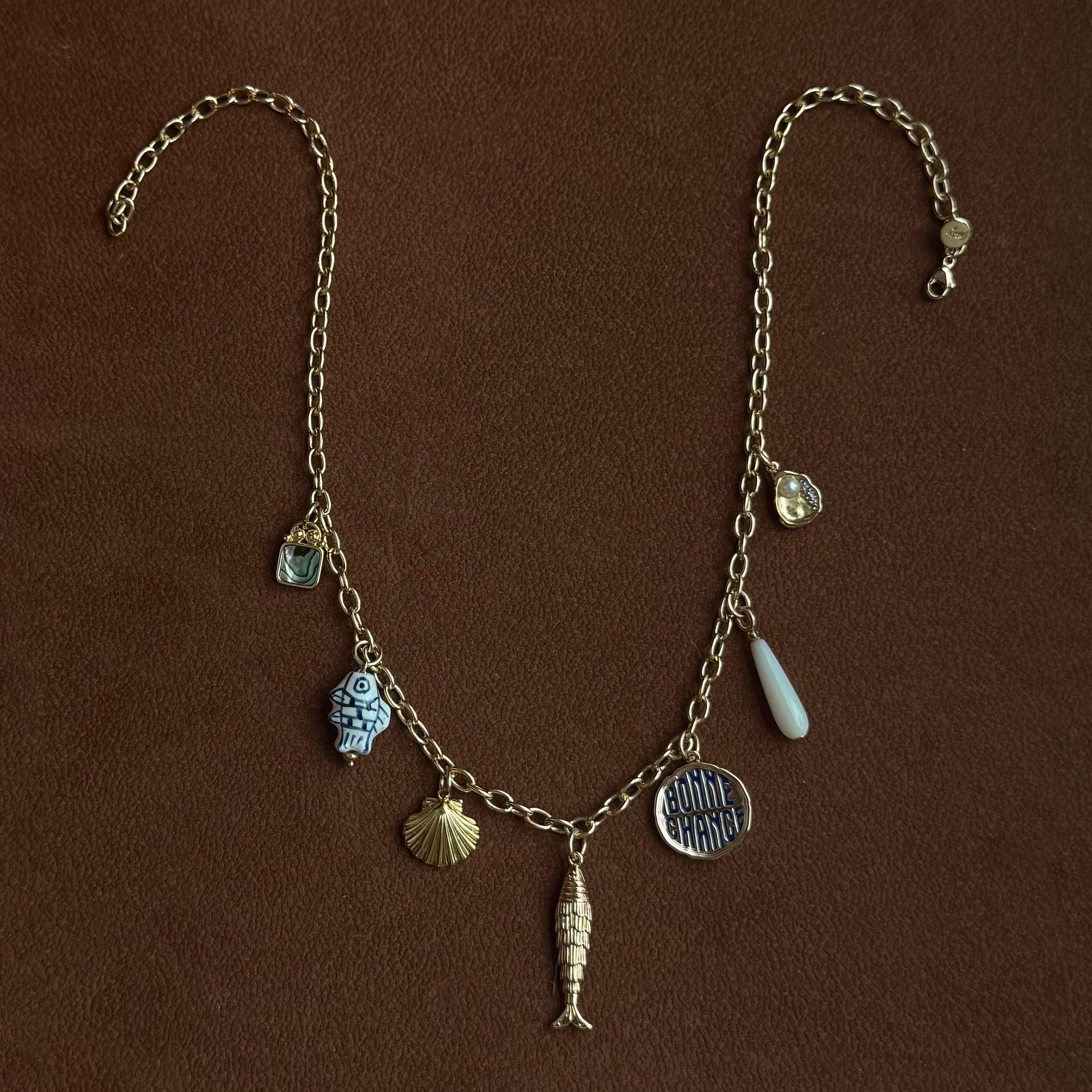 A gold chain necklace with various charms, including a fish, seashell, circular “Bonne Chance” pendant, blue face, clear stone, white teardrop, and blue gem, displayed on a brown surface.